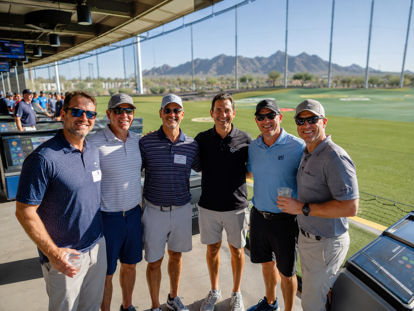 Crowd in casual polos standing in shaded Topgolf hitting bays overlooking a sunlit driving range