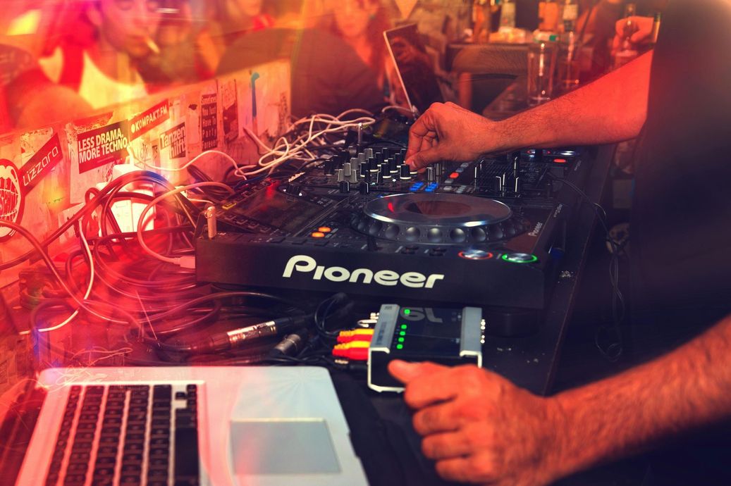 A DJ uses a Pioneer mixing console and a laptop to perform in a dimly lit, red-toned club setting.