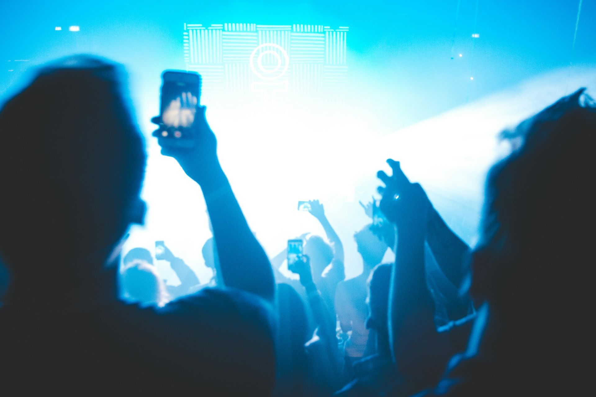 Silhouetted concert-goers with arms raised, holding phones up to record under bright blue stage lighting.
