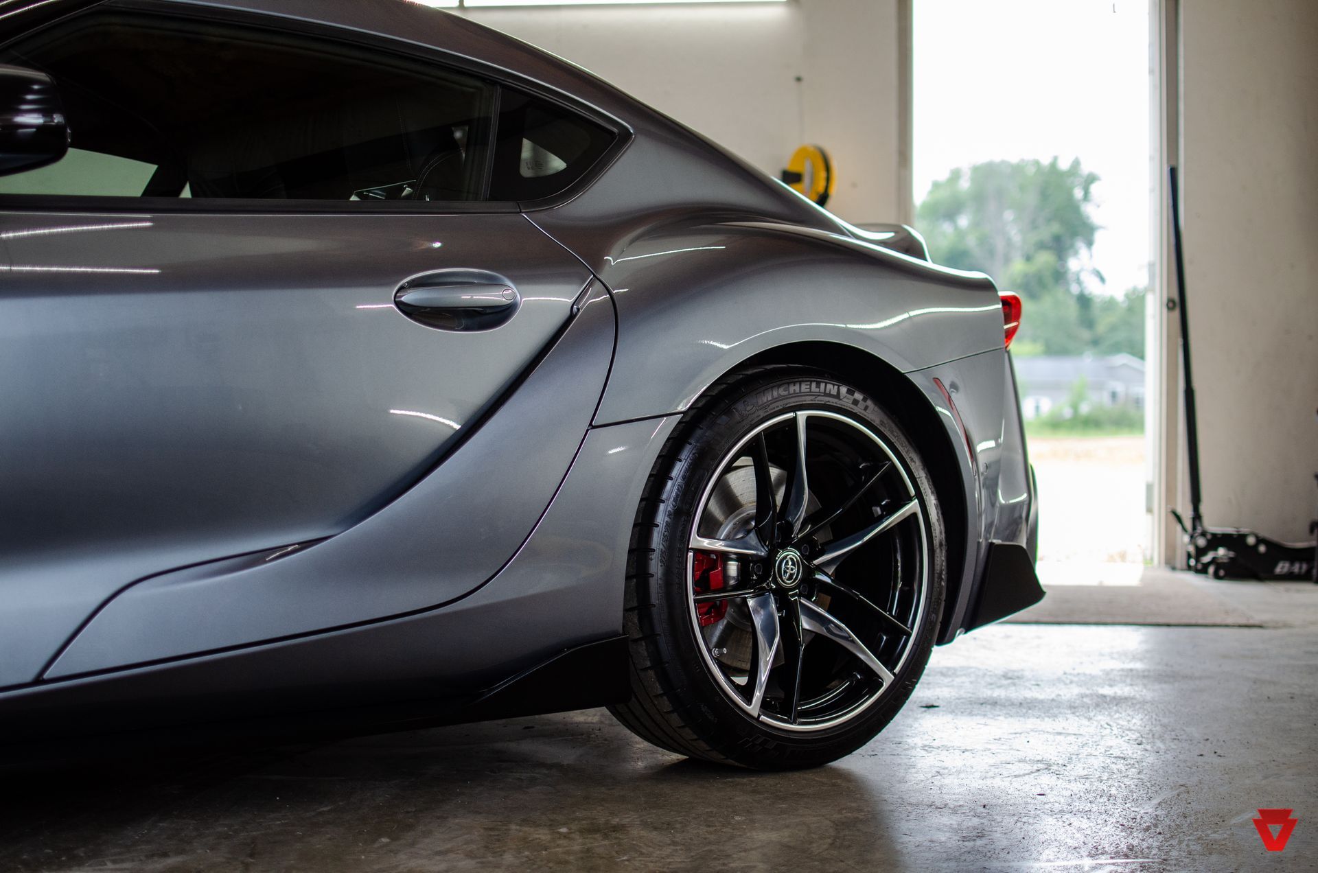 Gray sports car rear wheel and side panel in a garage, with red brake caliper and wet floor