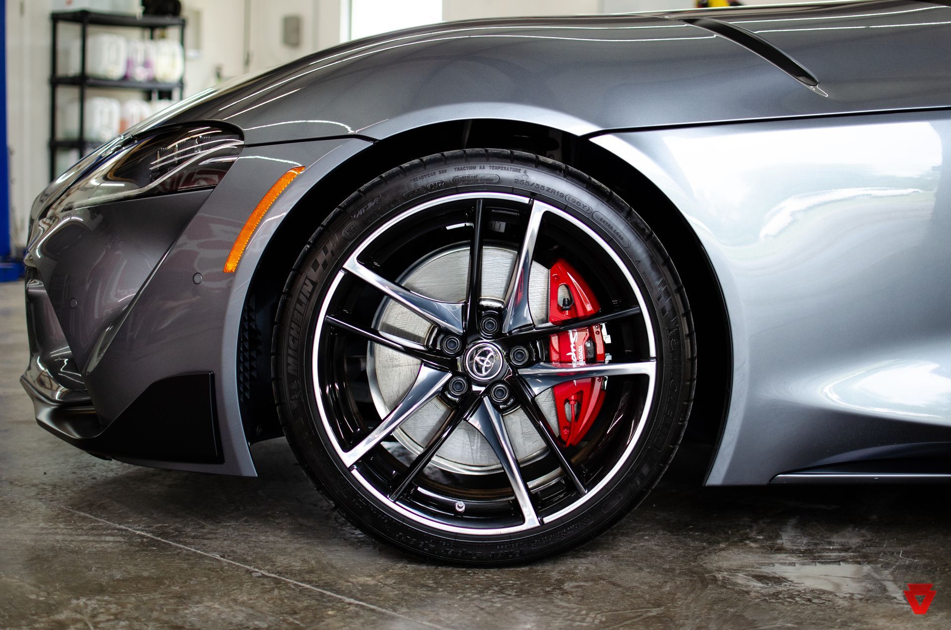 Close-up of a gray sports car front wheel with black rim and red brake caliper in a garage