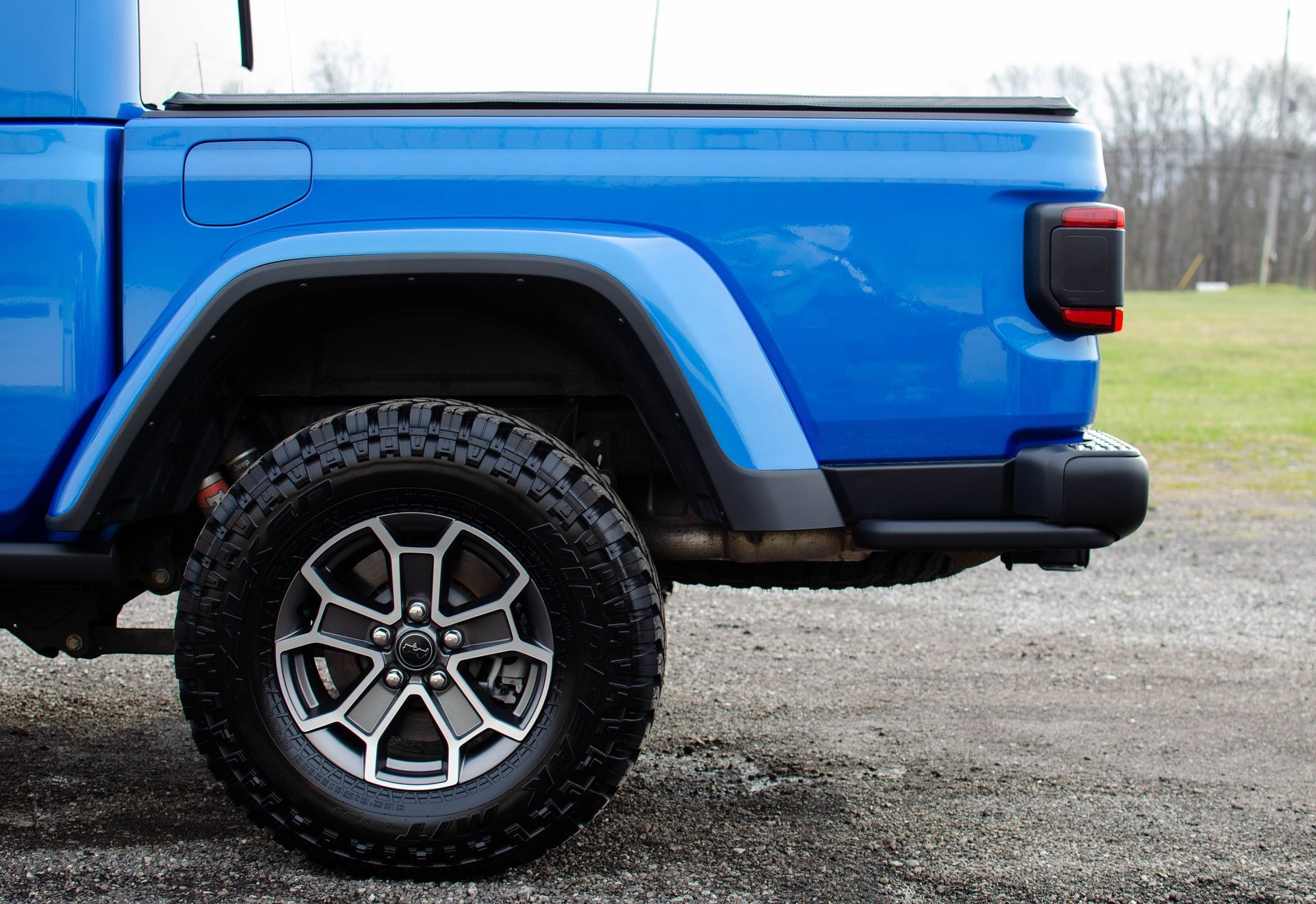 Blue pickup truck side view with off-road tire on gravel road, grassy field in background