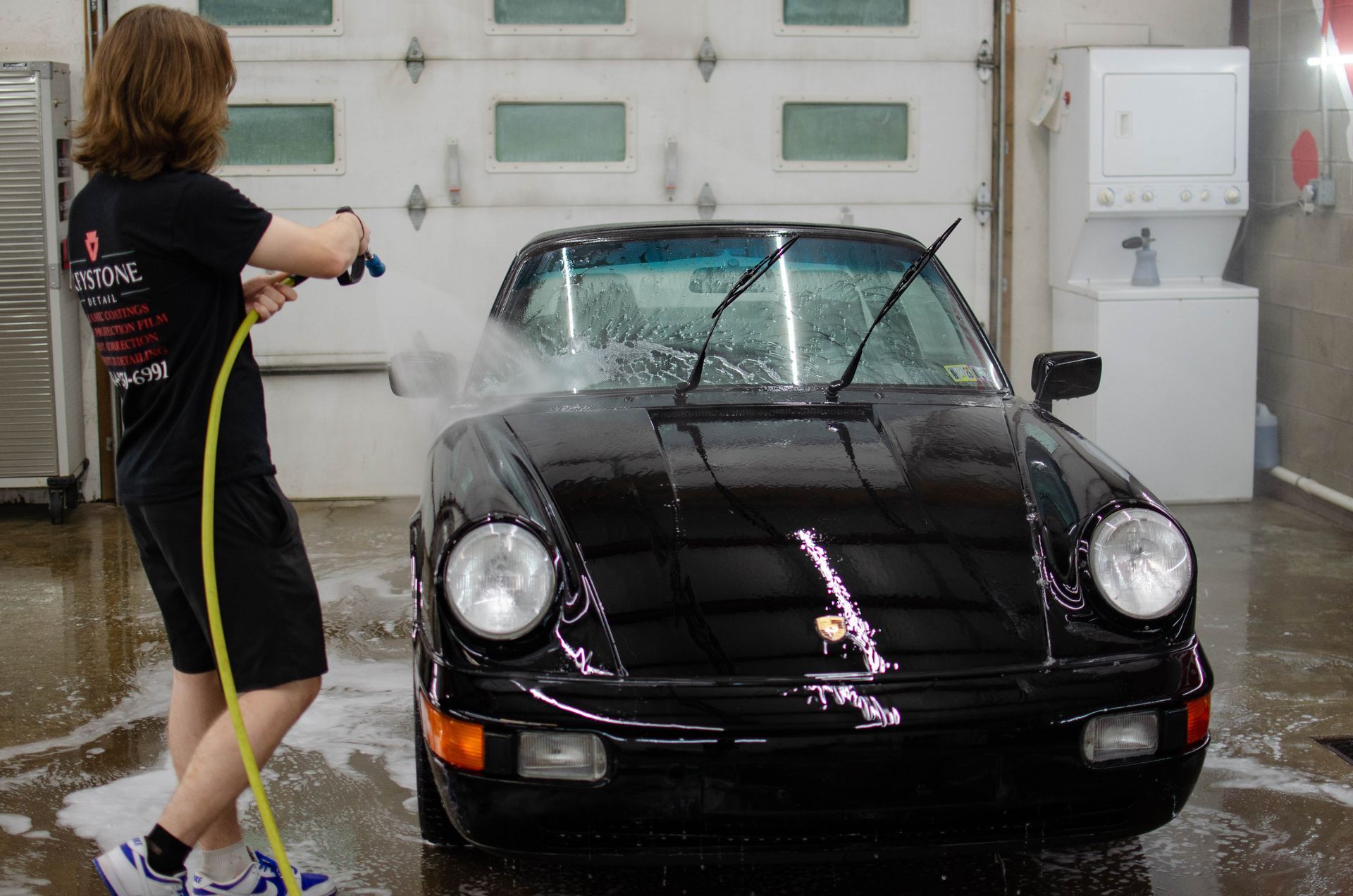 Person washing a black convertible sports car in a wet garage with a hose