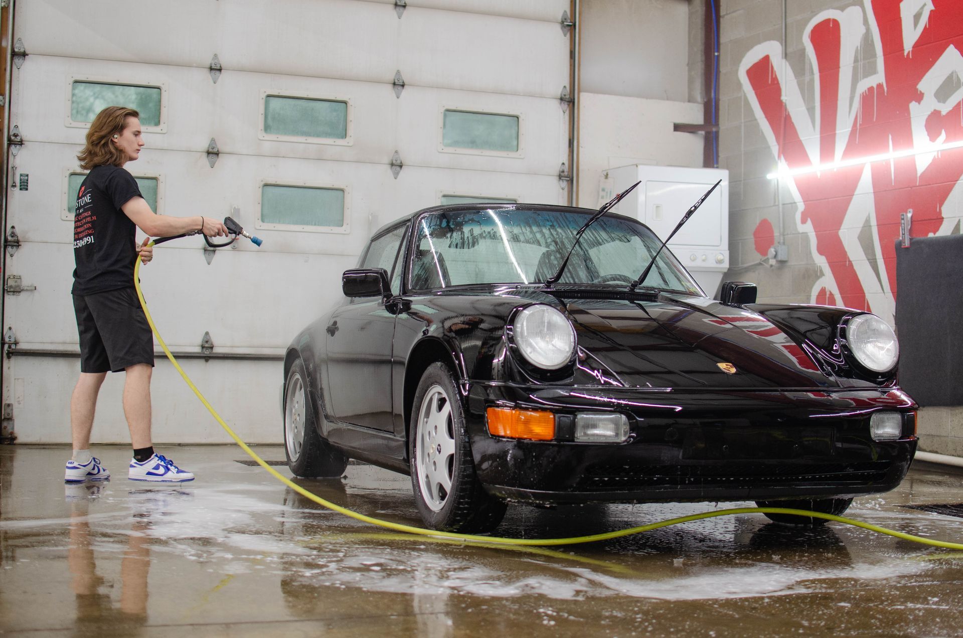 Person washing a black Porsche with a hose in a wet garage, spray hitting the car.