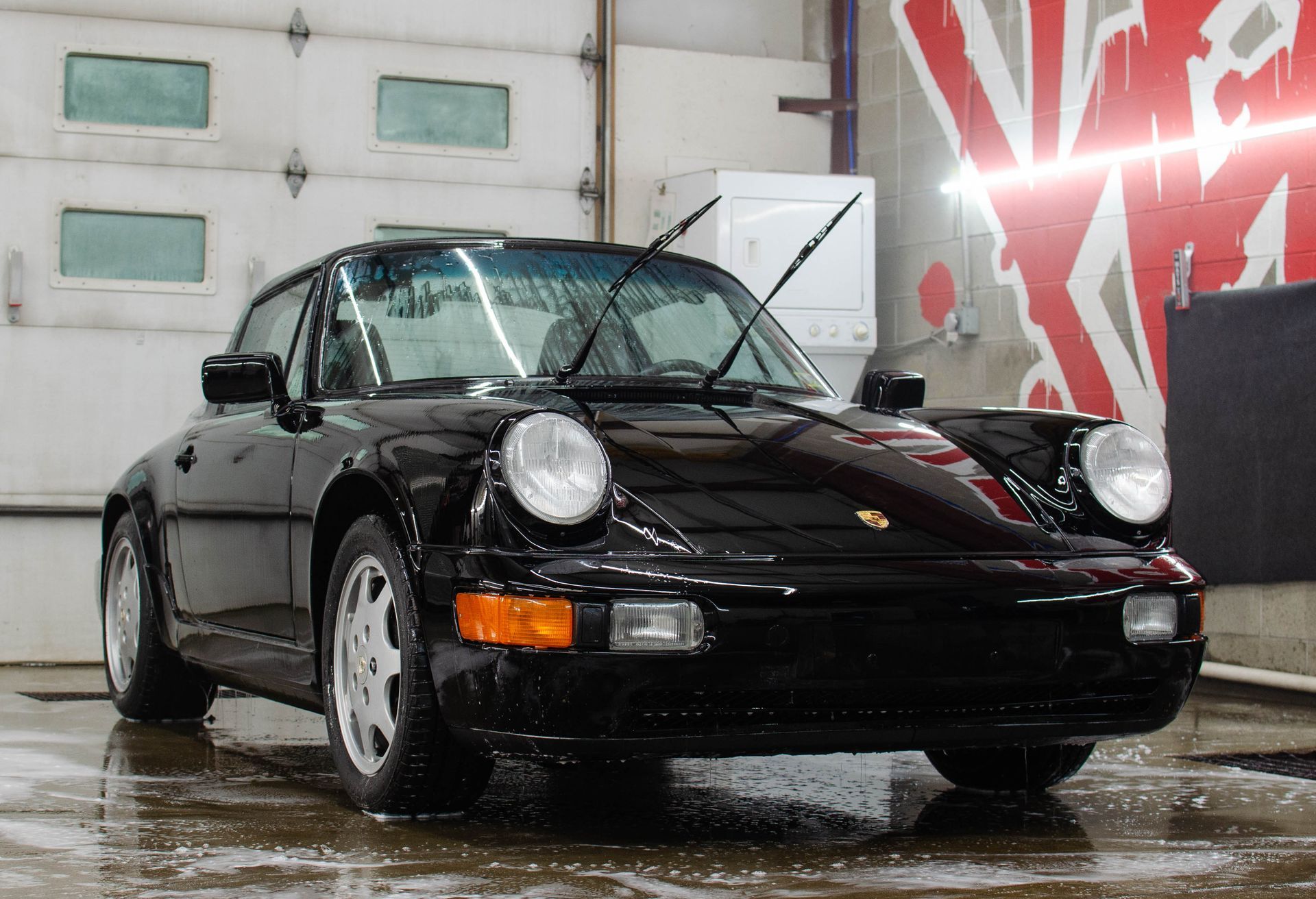 Black Porsche 911 parked on a wet floor in a garage, front view with headlights on and red mural background