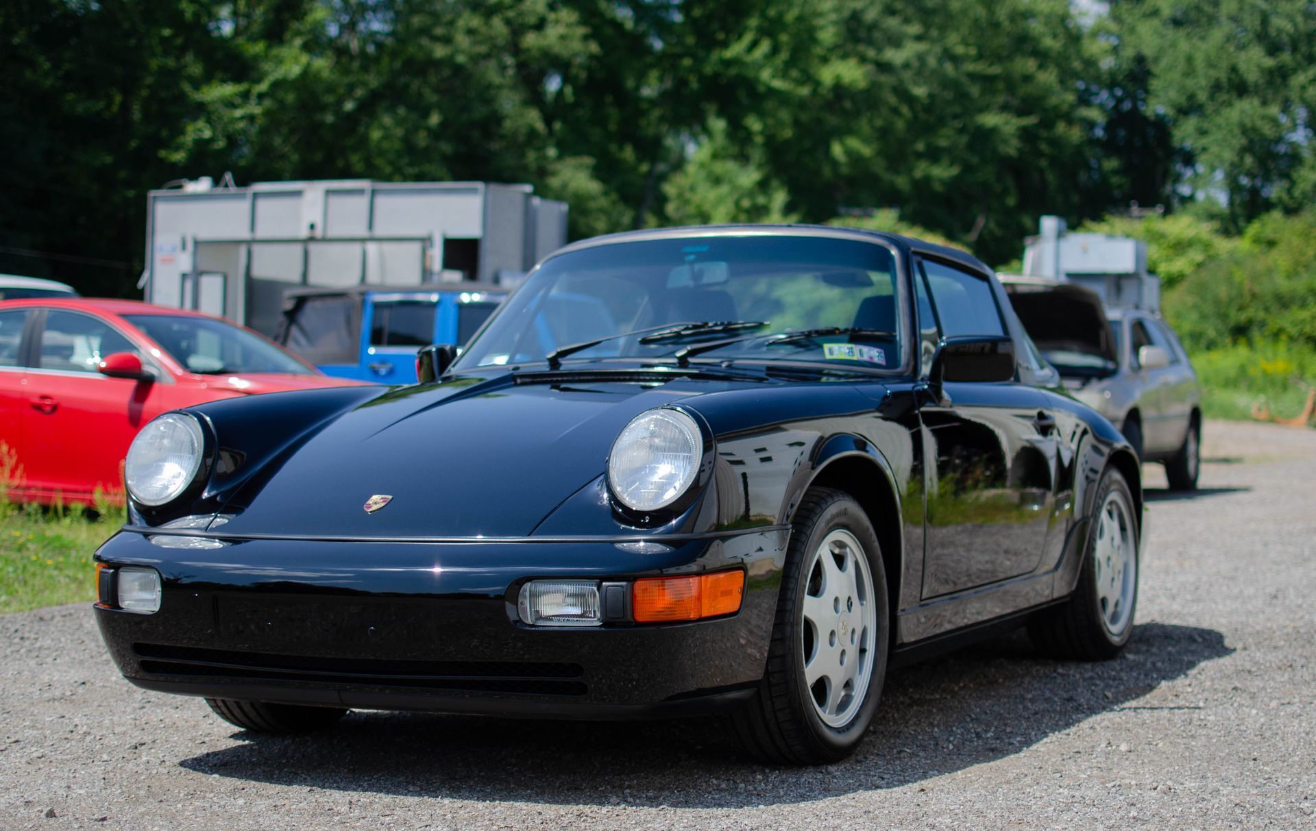 Black Porsche 911 parked outdoors on a gravel lot, with other cars and buildings in the background