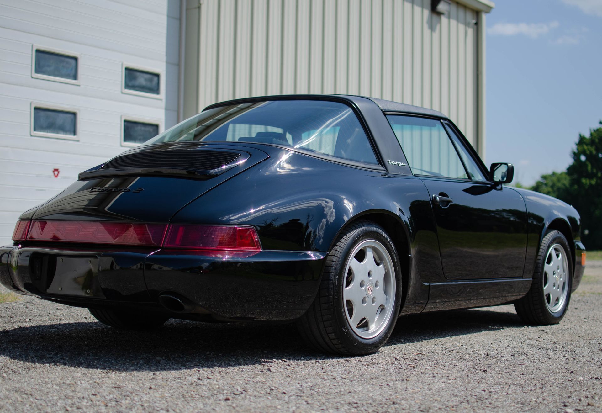 Dark blue classic Porsche 911 parked beside a light industrial building on gravel.
