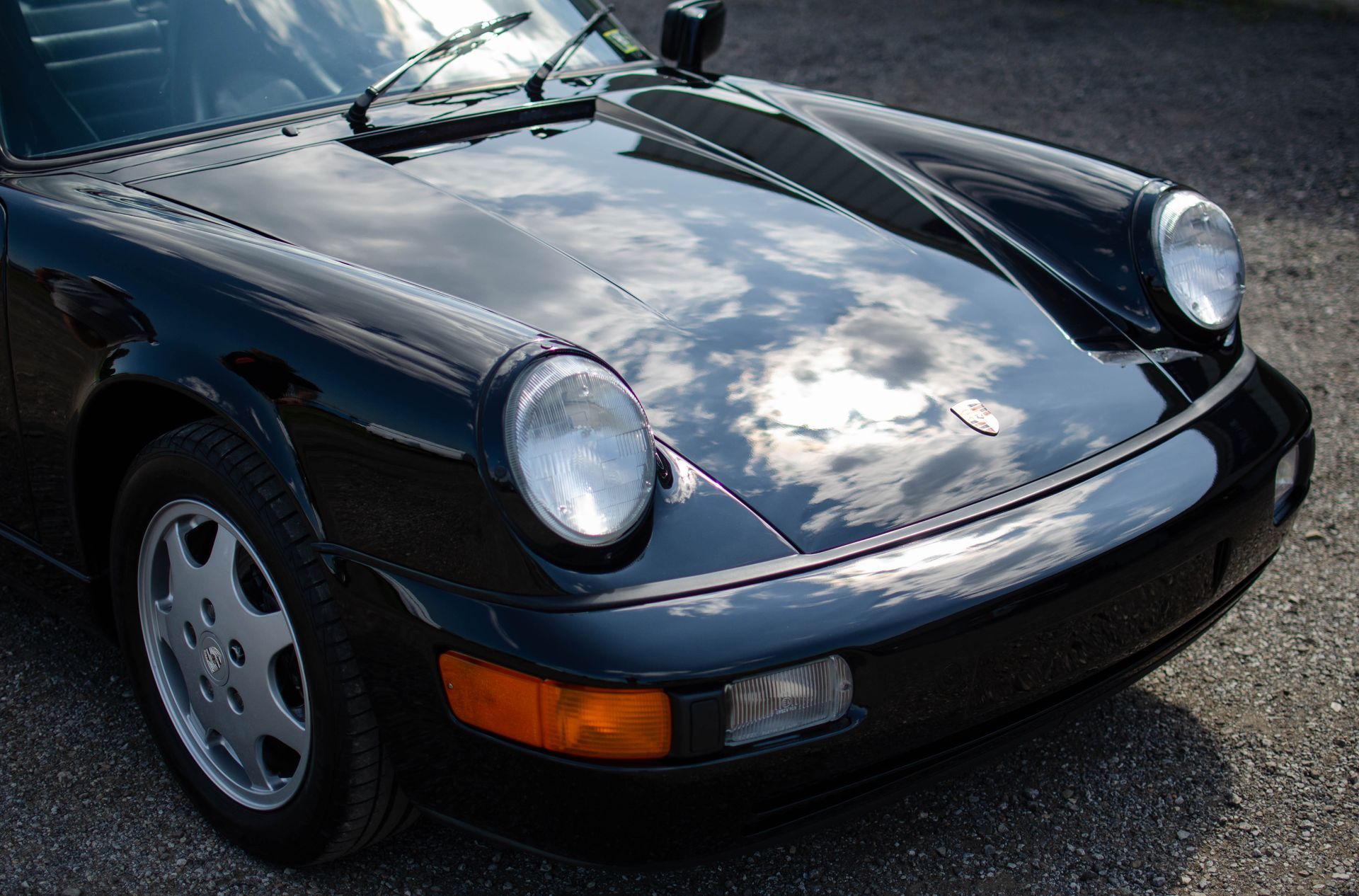 Close-up of a black sports car’s front end parked on pavement, with sky reflections on the hood.