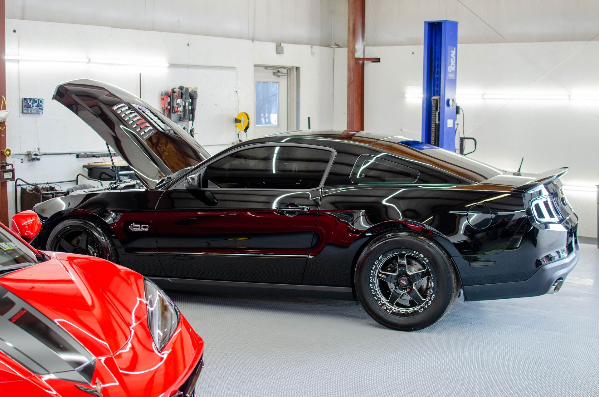Black sports car in a garage with hood open; red car partially visible in the foreground.