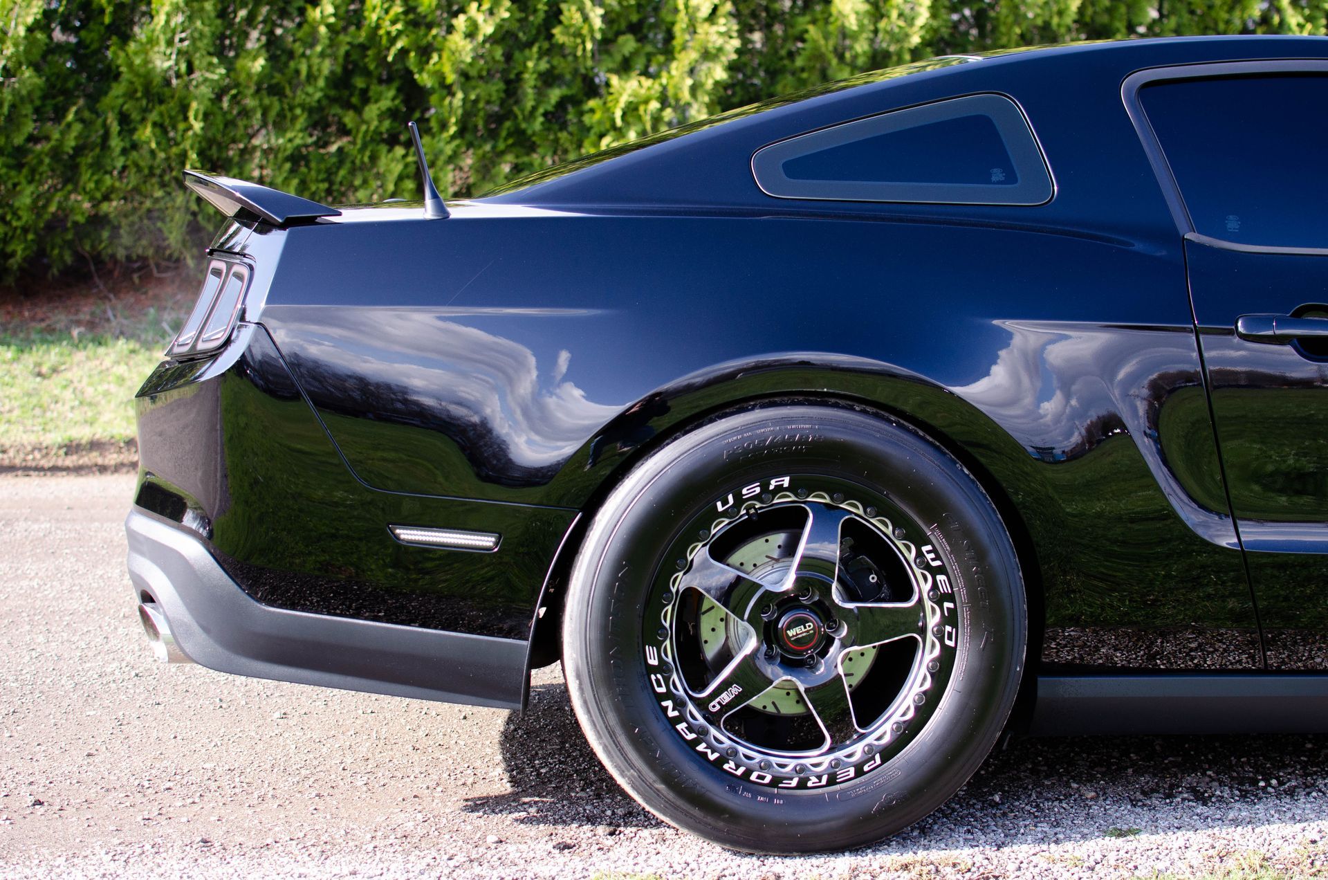 Dark blue sports car rear side with black rims parked on gravel near greenery
