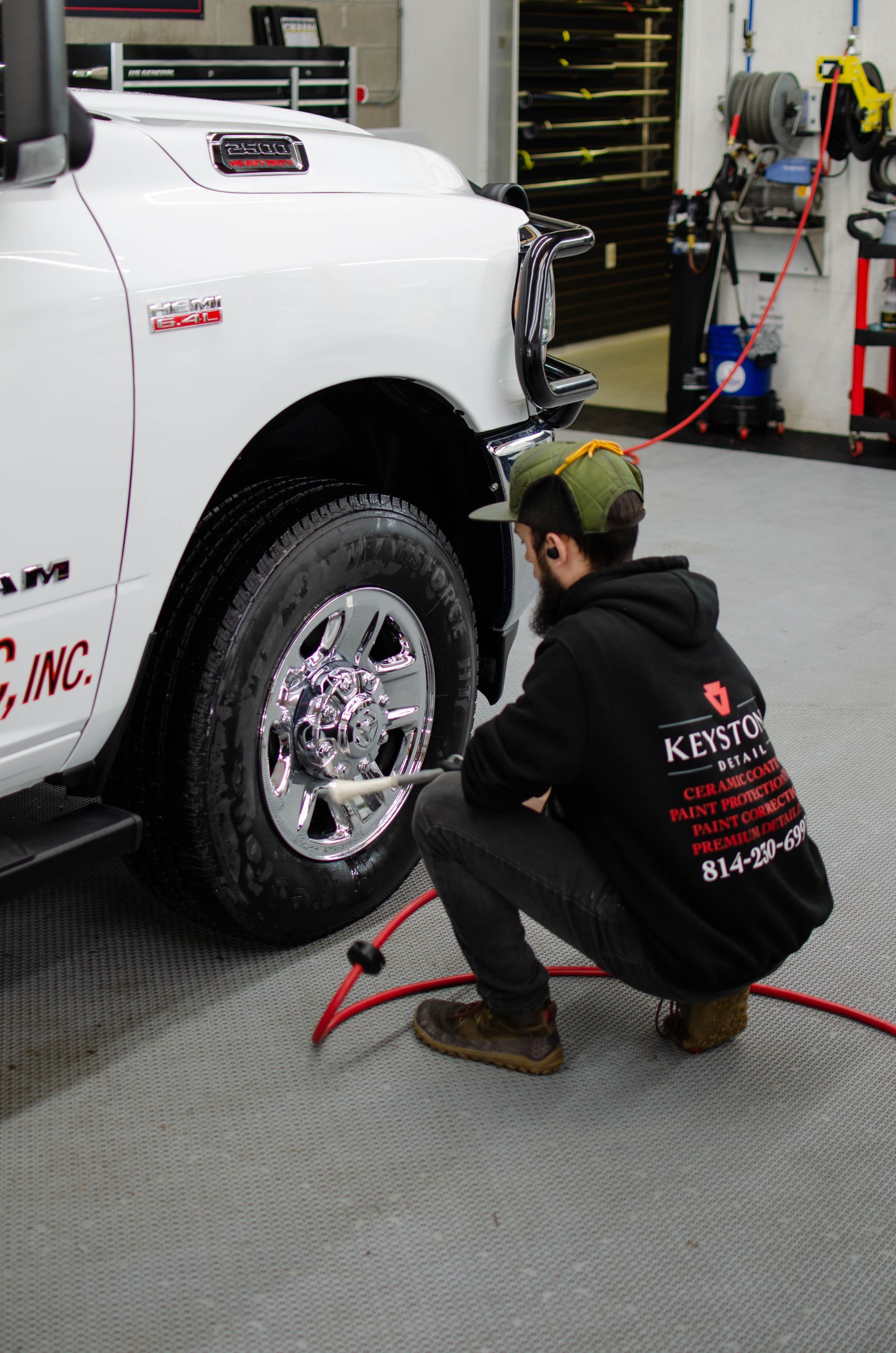 Mechanic kneels by a white pickup truck’s wheel in a garage, checking tire pressure with a red hose.