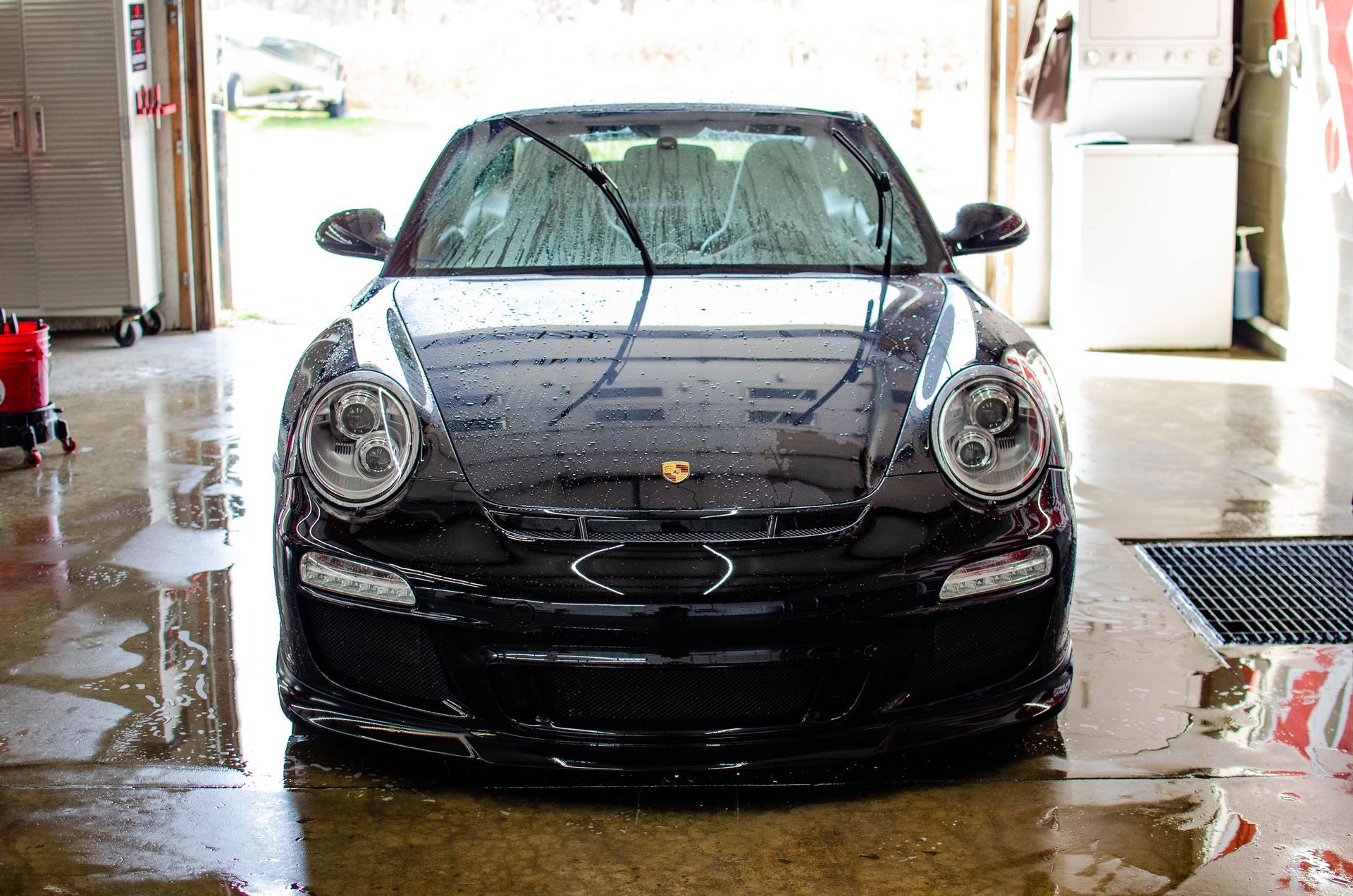 Black sports car being washed in a garage, covered in soap and water with wet floor reflections