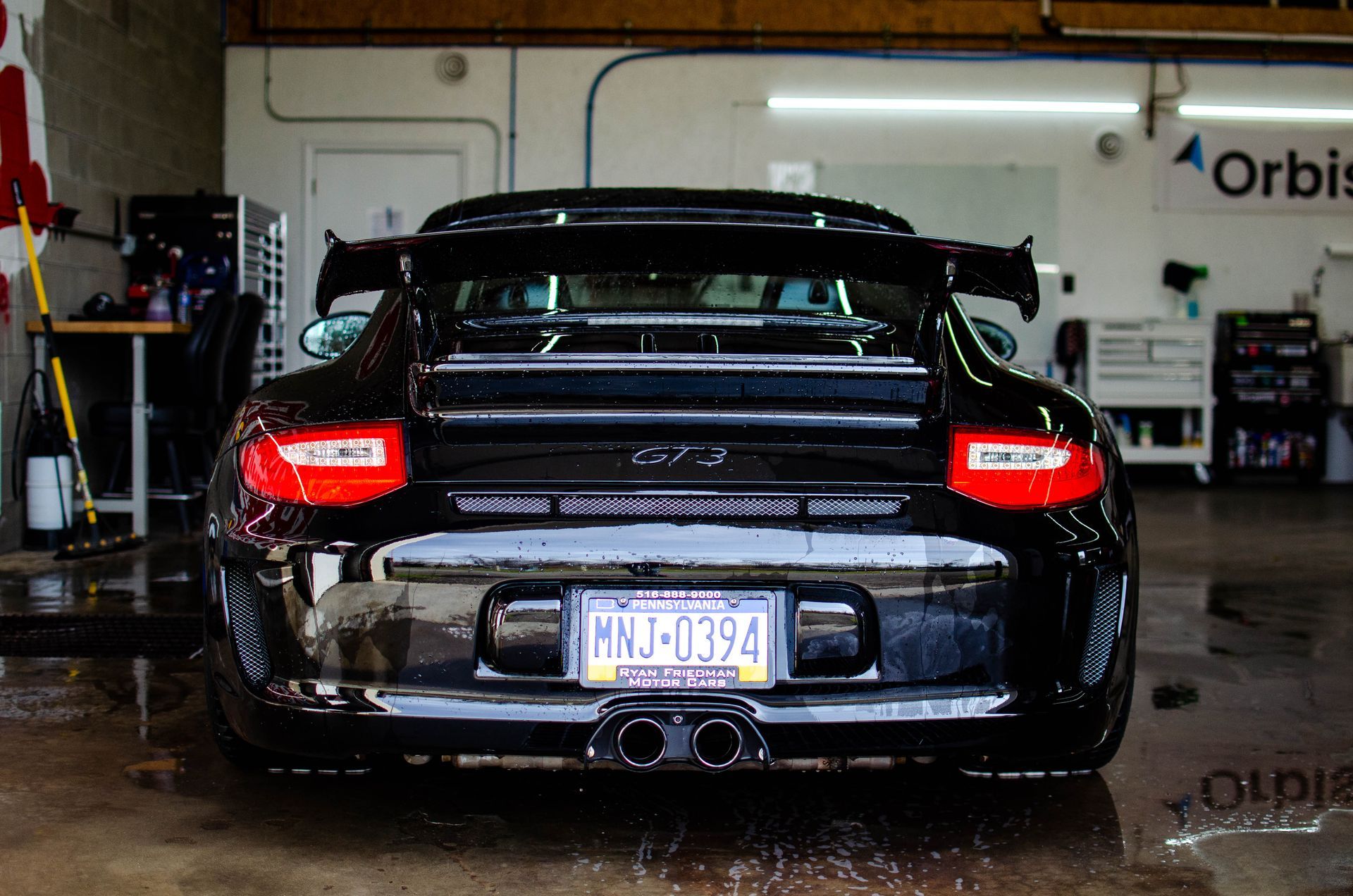 Rear view of a black sports car in a garage, with taillights on and a visible license plate.
