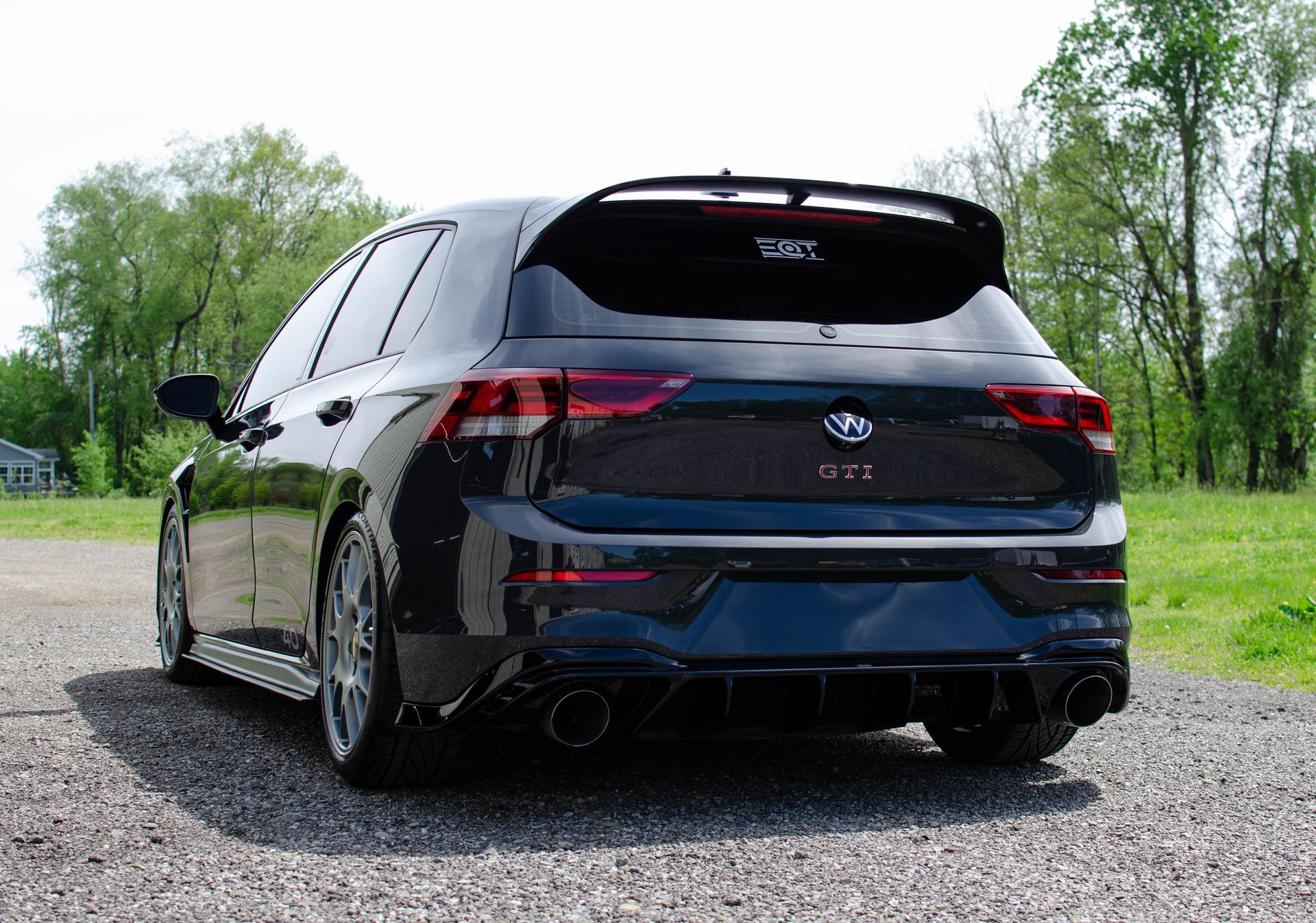 Black hatchback car parked on gravel, rear view, with trees in the background