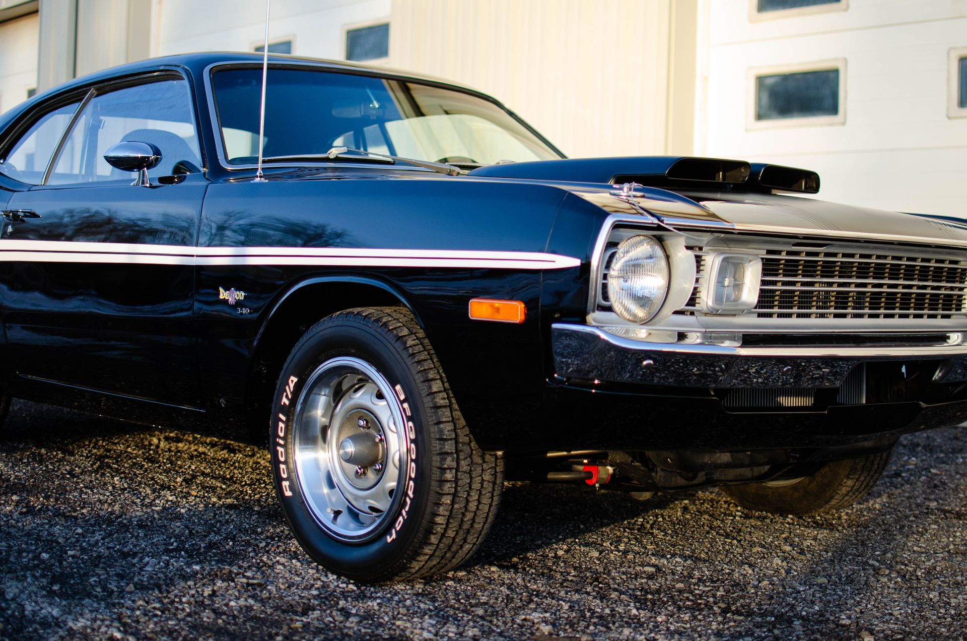 Black classic muscle car parked on gravel, front-left view, with chrome trim and hood scoop