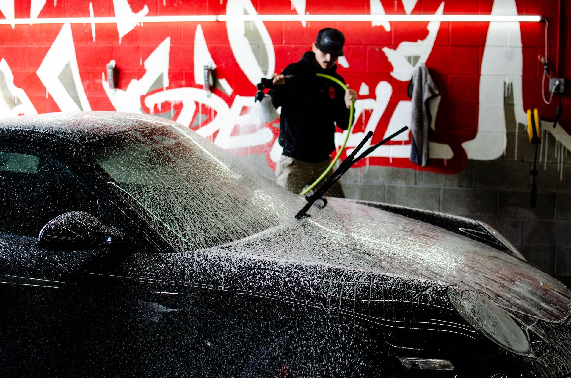Person pressure-washing a black car in front of a red graffiti wall