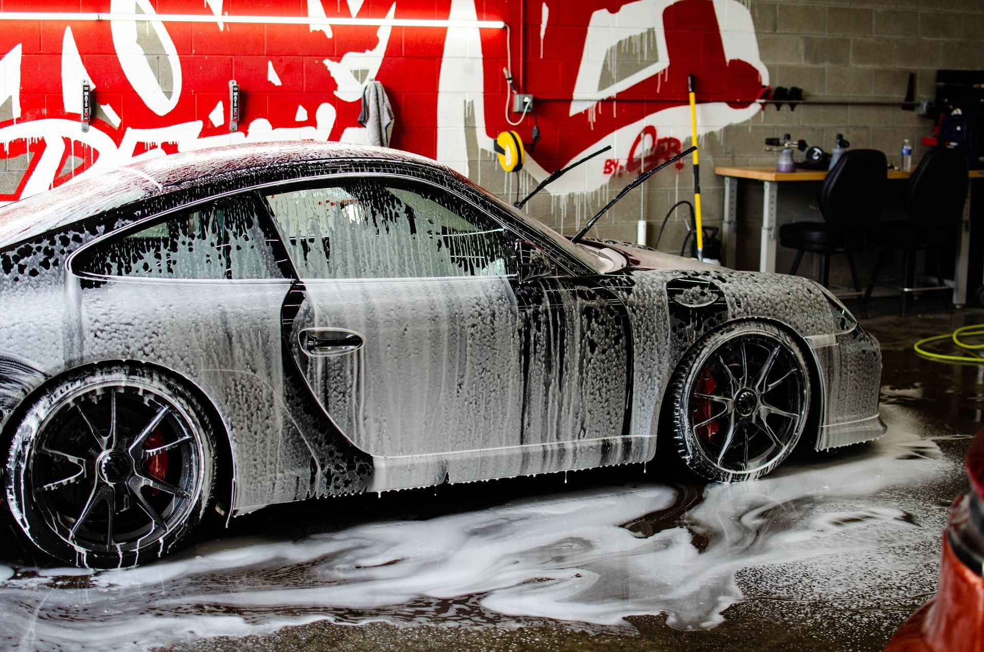 Silver sports car being hand-washed in a garage, covered in white soap suds.