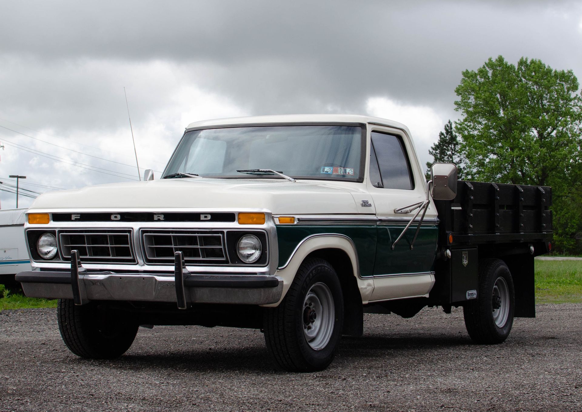 White and green pickup truck with black flatbed parked on gravel under a cloudy sky