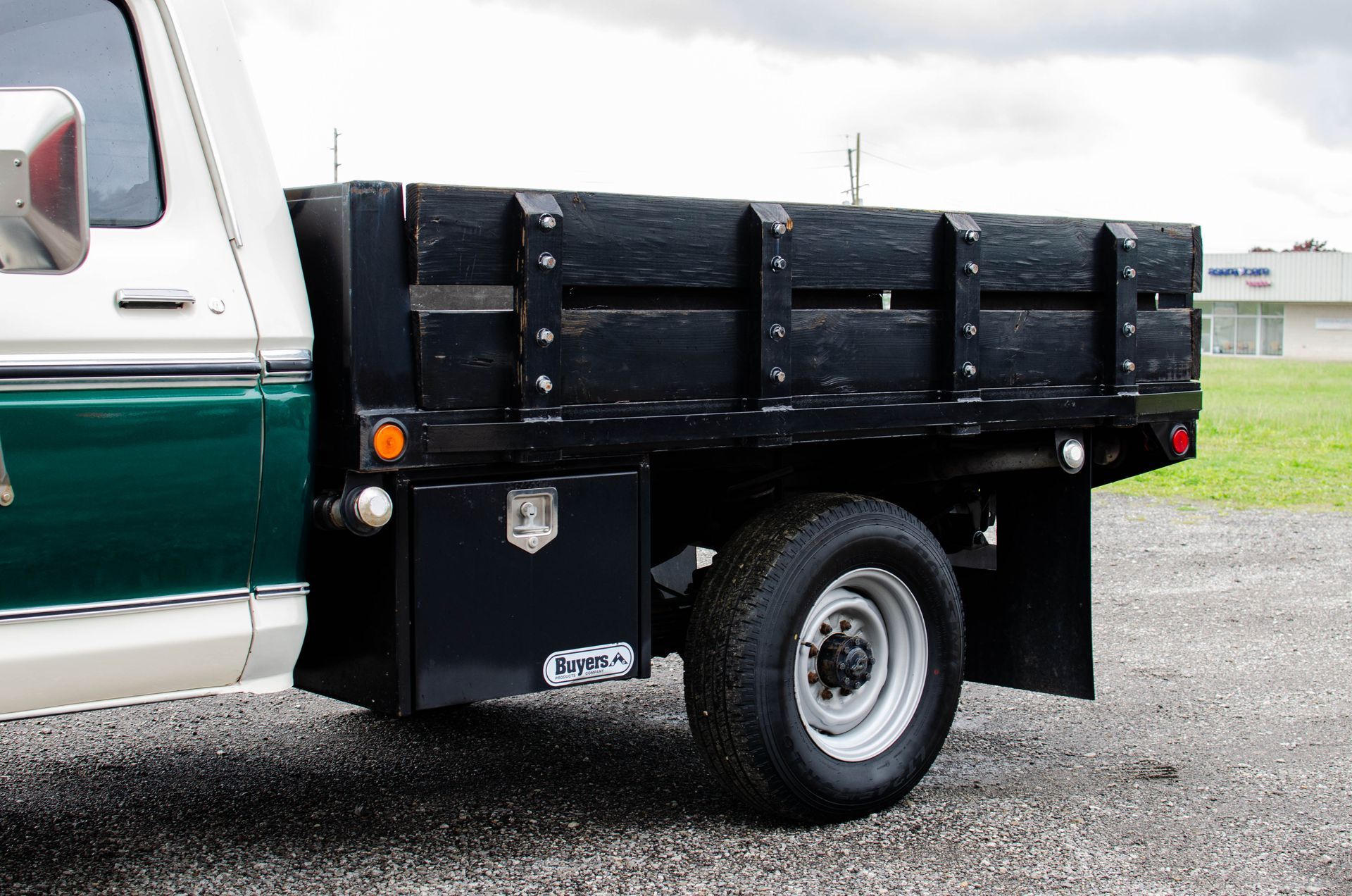 Black flatbed truck bed on a white and green pickup, parked on gravel outdoors.