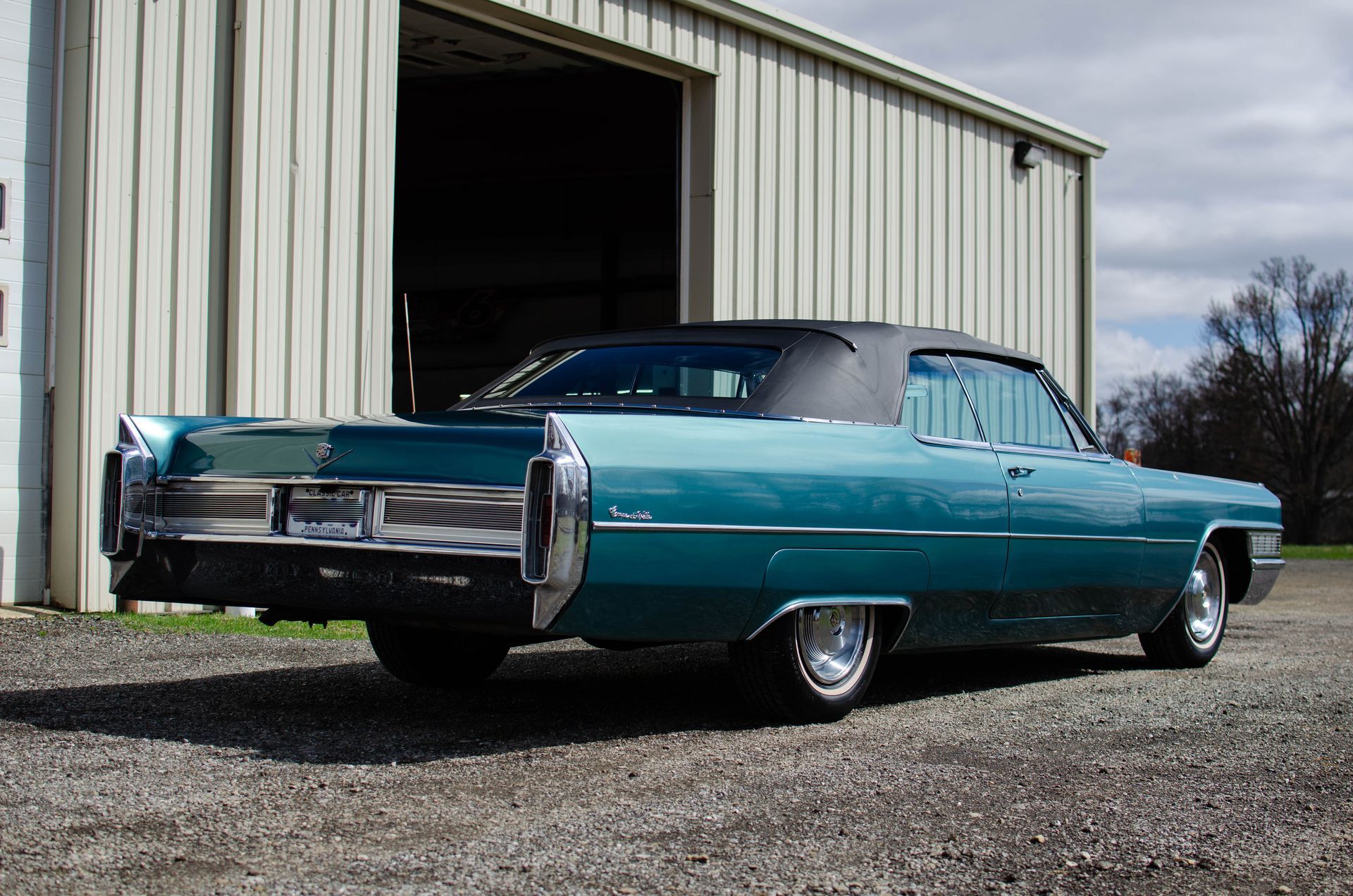 Turquoise classic car parked outside a metal garage on gravel.