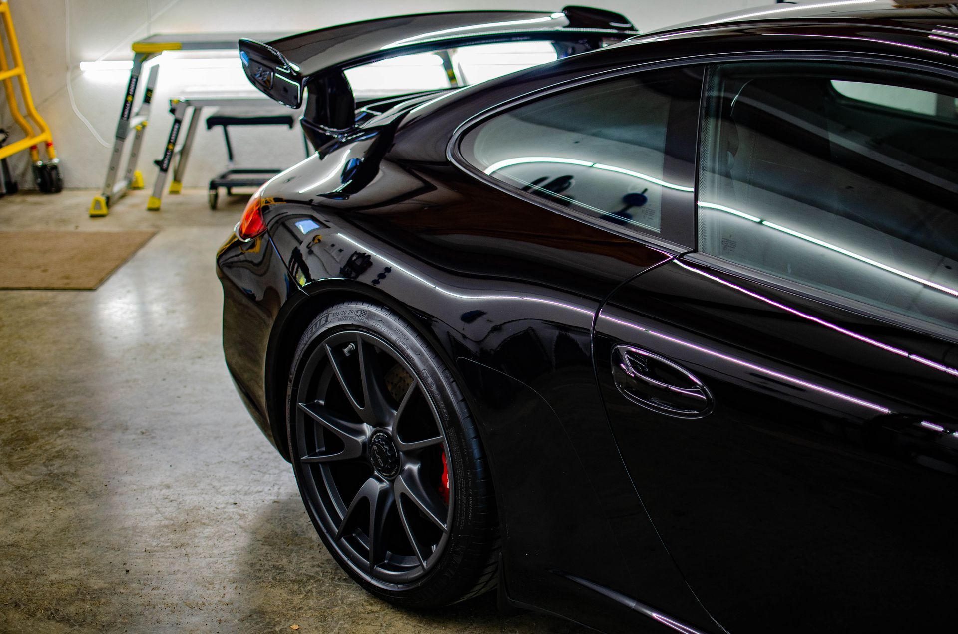 Black sports car parked in a garage, rear side view with raised spoiler and black rims