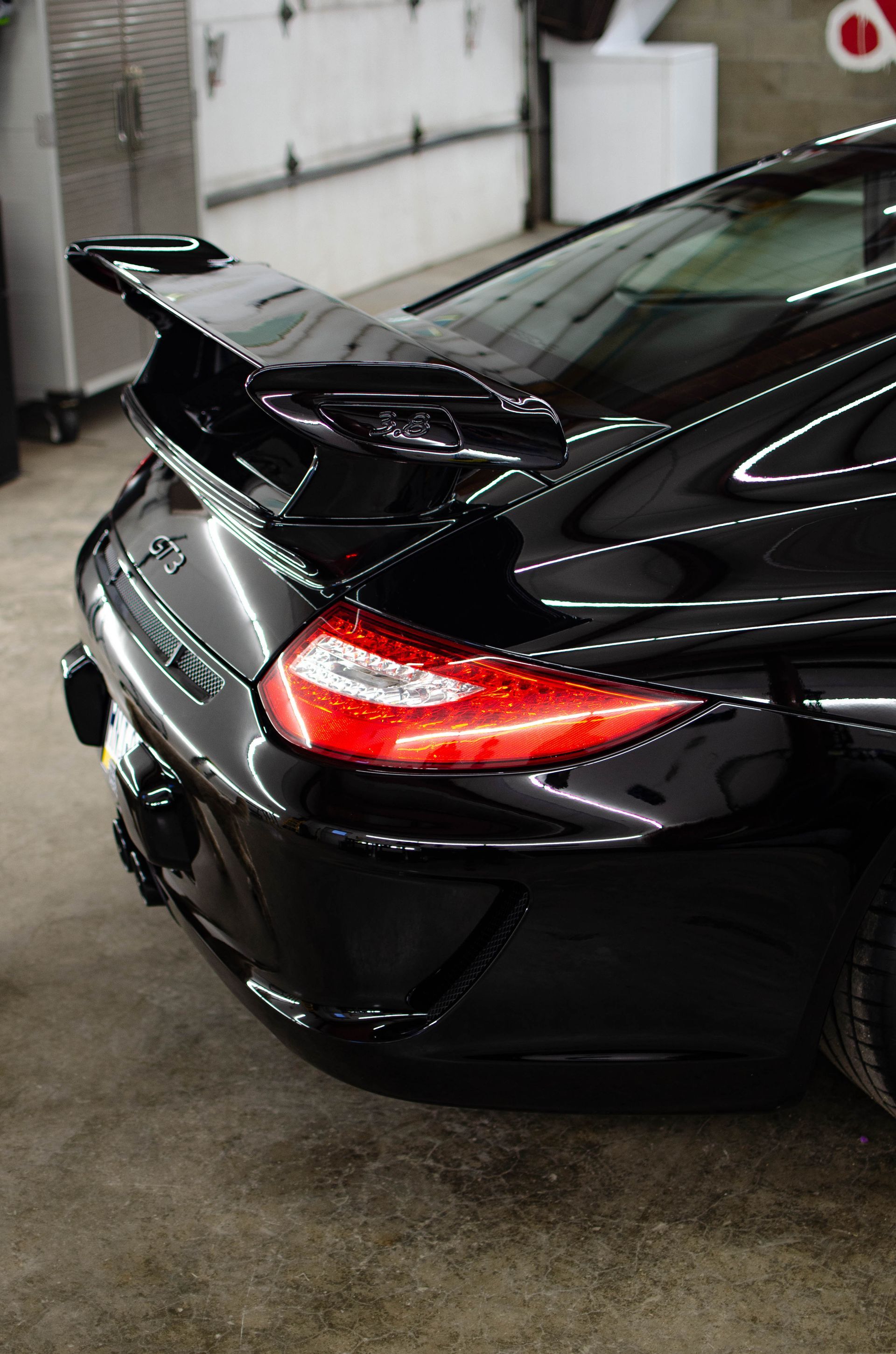 Black sports car rear in a garage, with a large rear spoiler and red taillight illuminated.