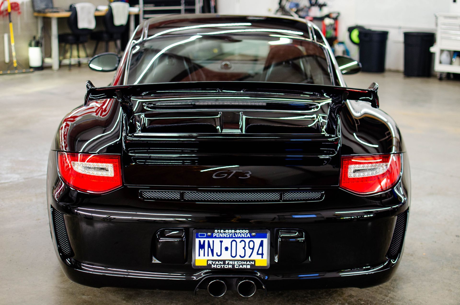 Rear view of a black sports car with a rear spoiler, parked indoors with red taillights and a blue license plate.