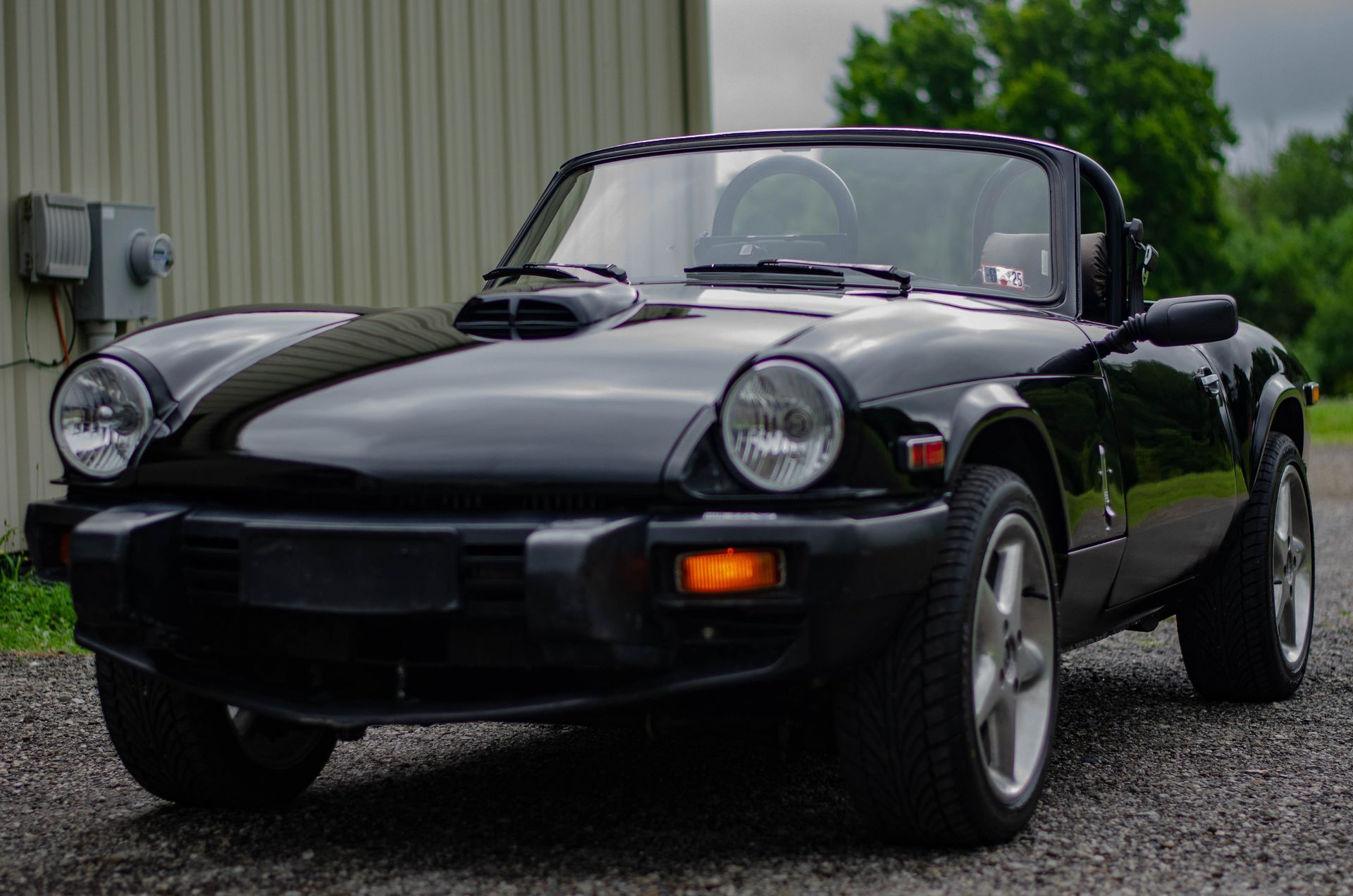 Black classic convertible sports car parked on gravel beside a metal building.