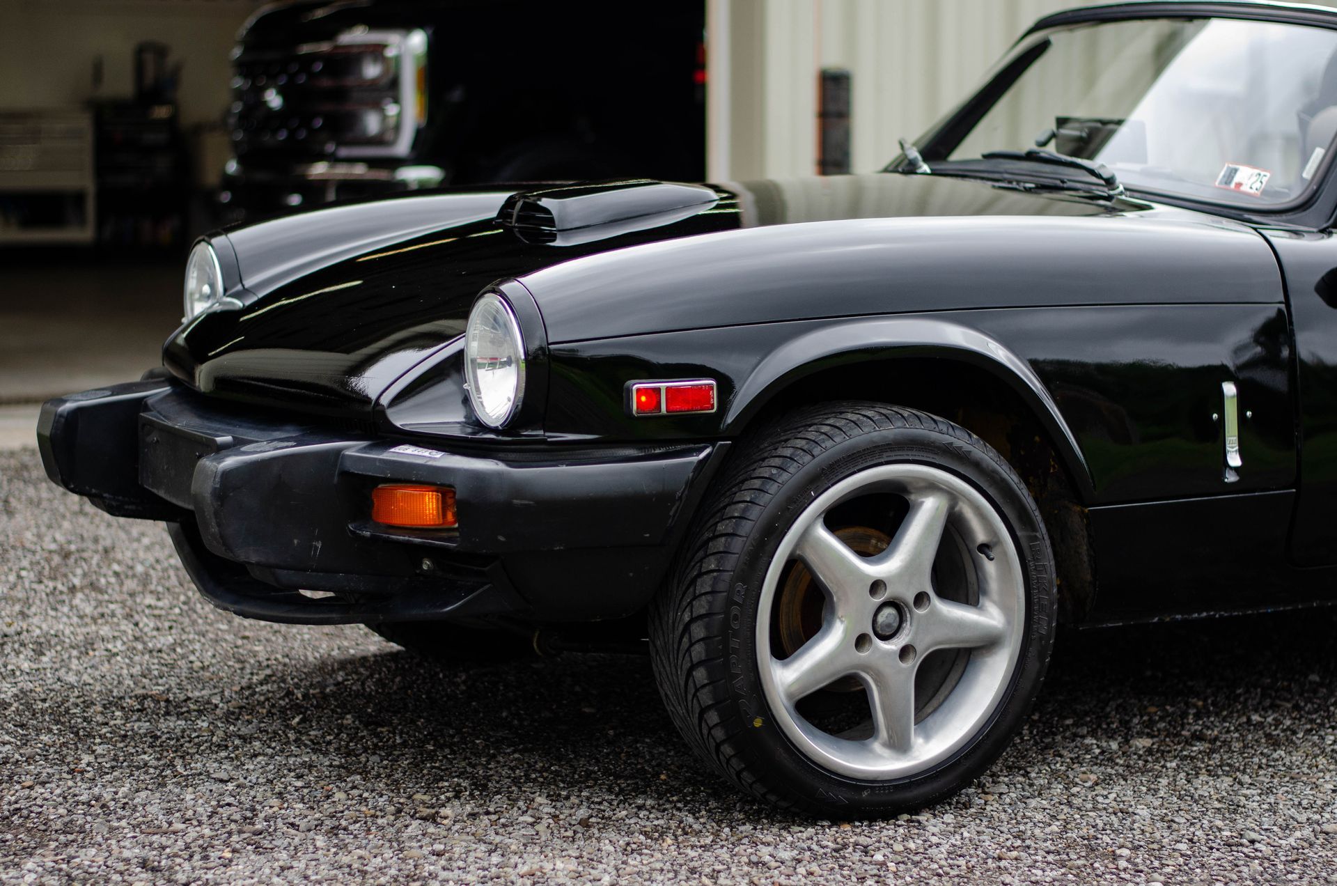 Black classic sports car parked on a gravel lot, front-left view, with silver five-spoke wheels