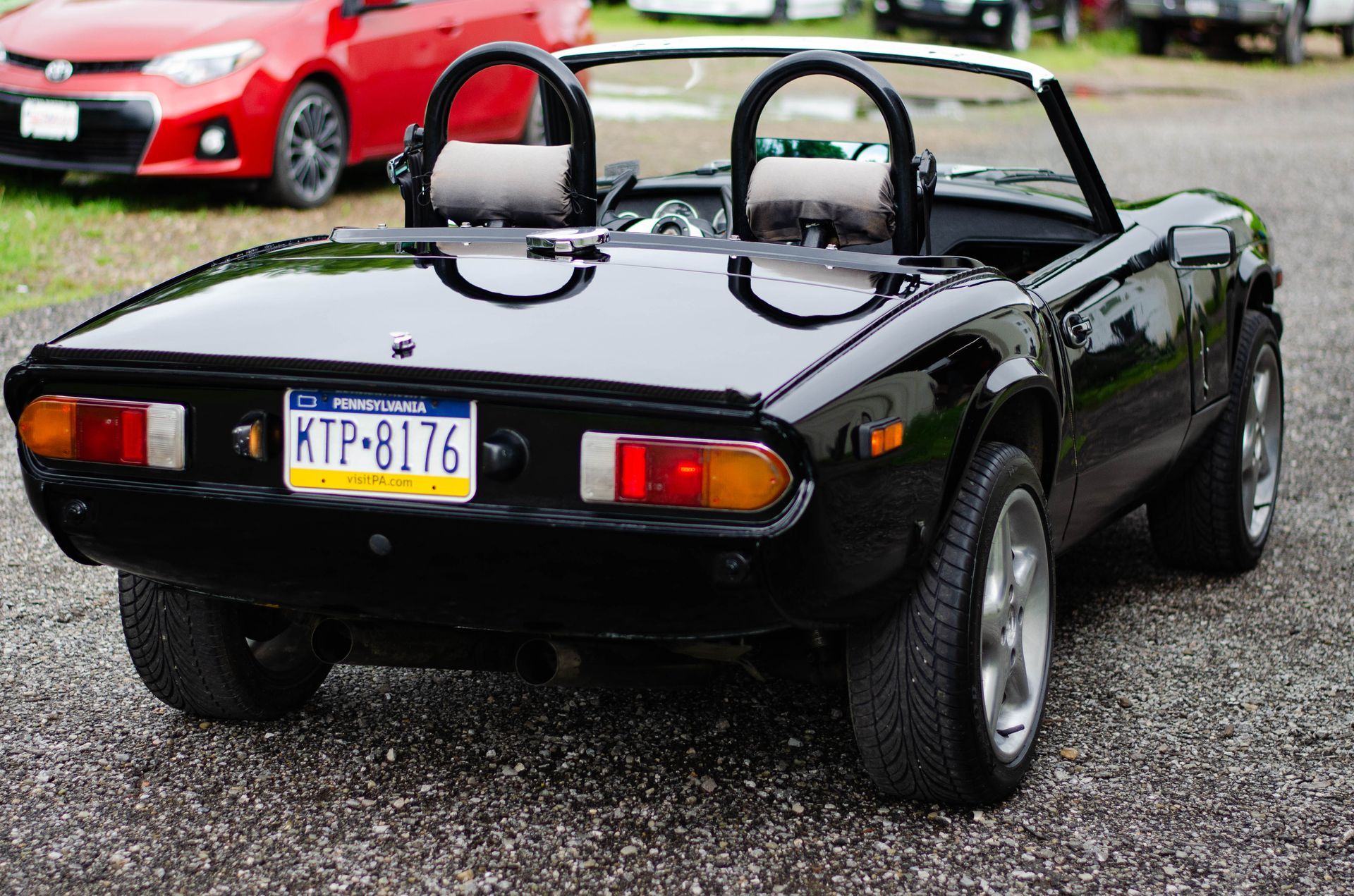 Black convertible sports car viewed from the rear on a gravel lot, with a red car in the background