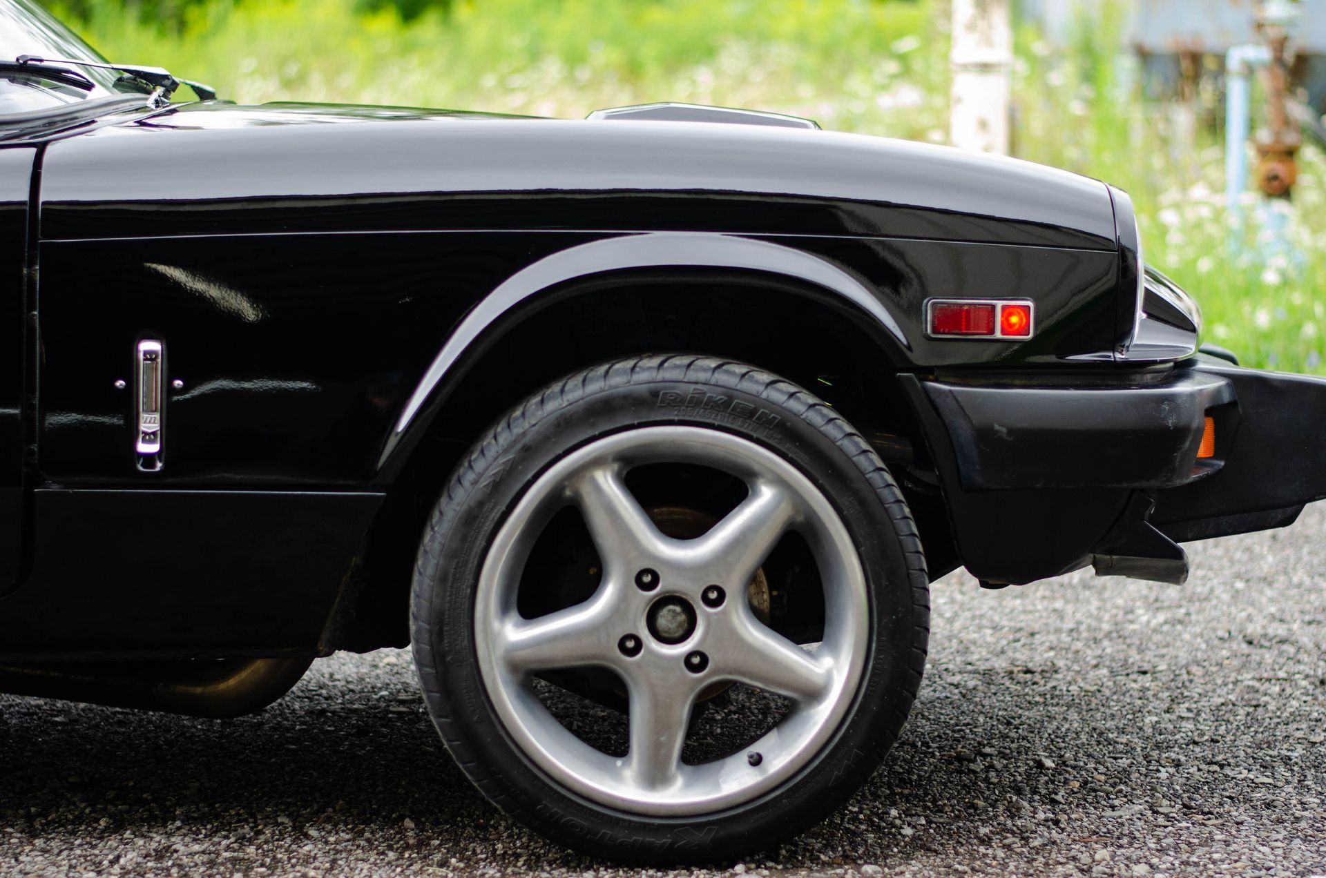 Black sports car front wheel and headlight on a gravel driveway, with a silver rim visible.