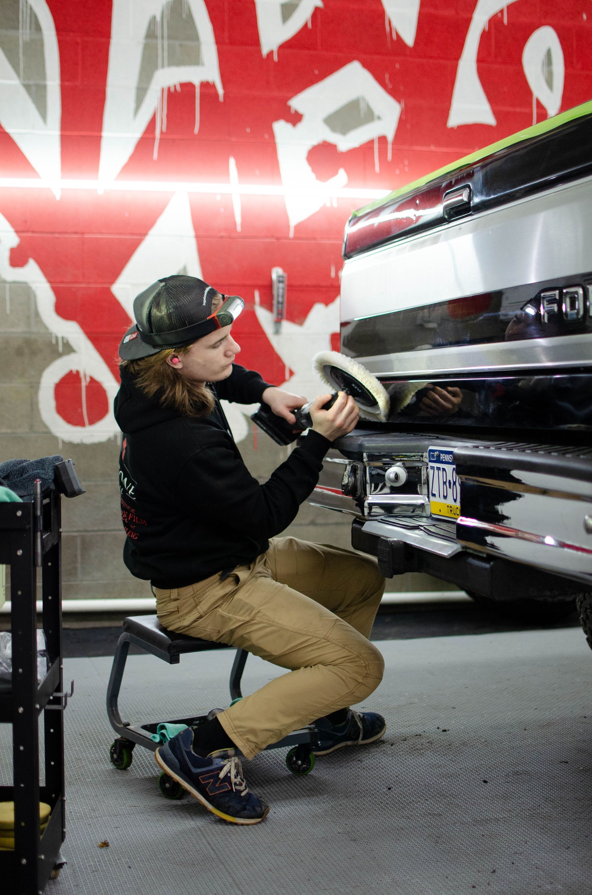 Person kneeling and polishing a car bumper in an auto detailing shop with red graffiti wall.