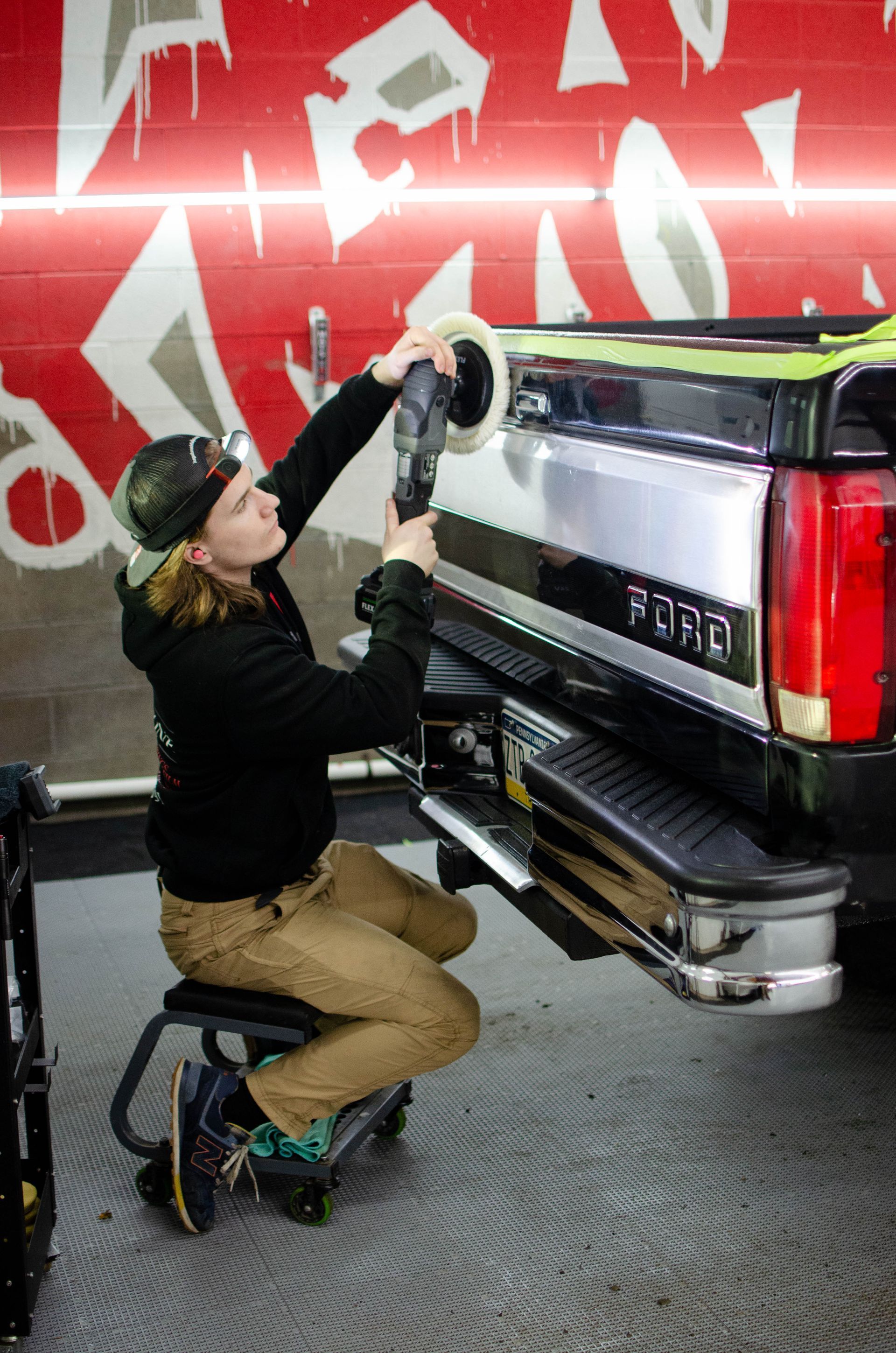 Mechanic polishing the rear of a black pickup truck in a garage, with a red graffiti wall behind.