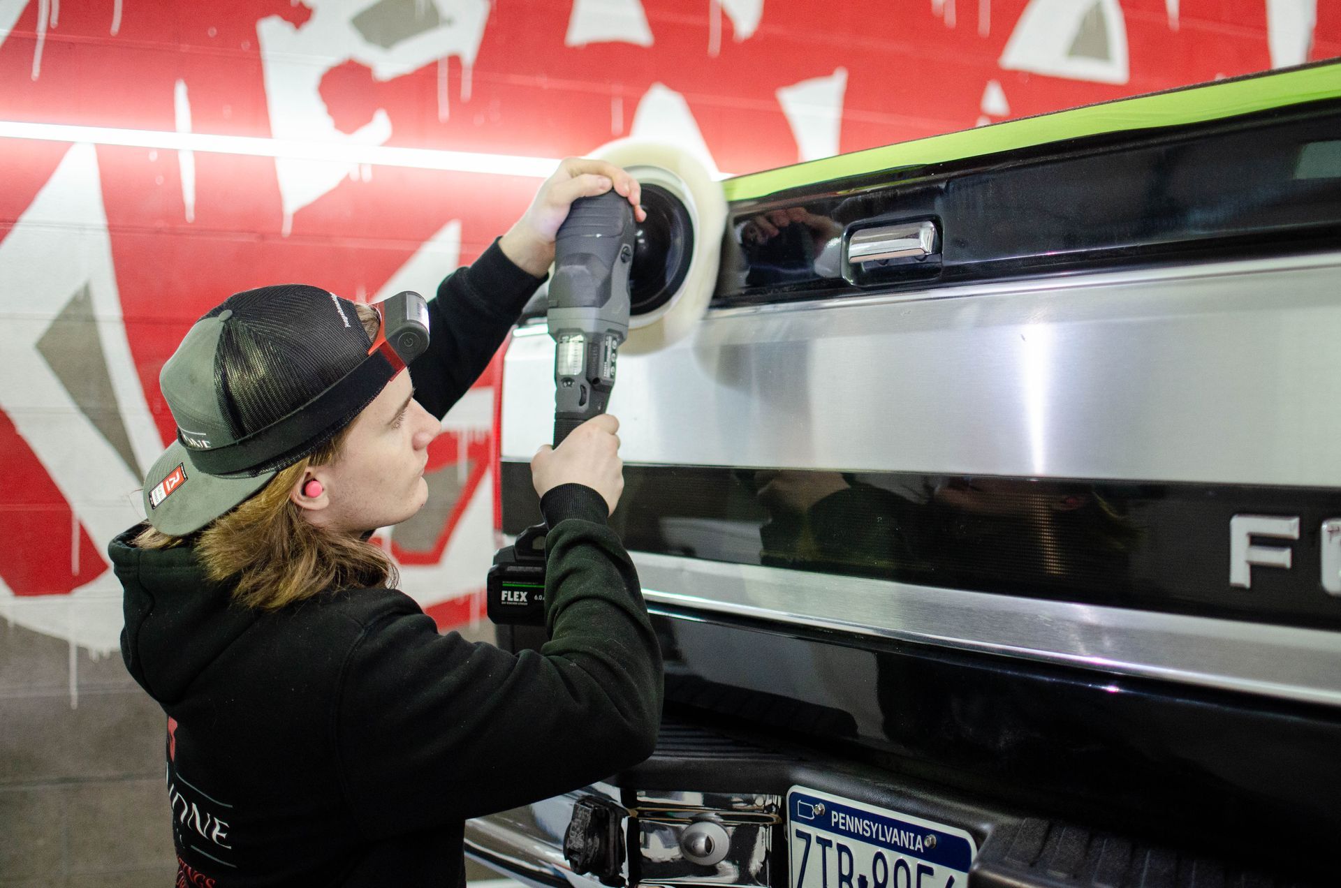 Person polishing a black car’s rear with a buffer in a workshop, red wall in the background
