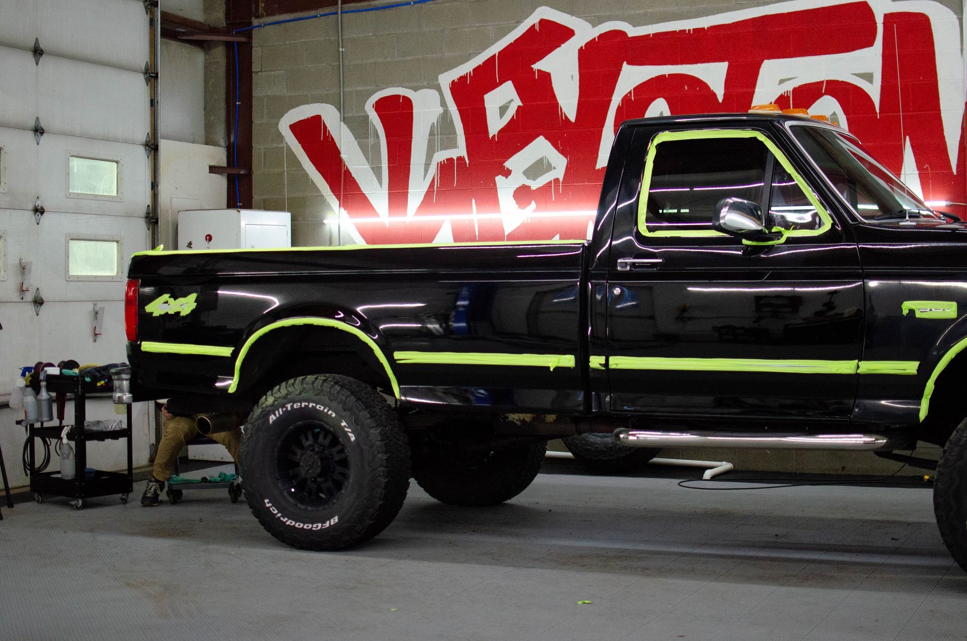 Black lifted pickup truck with neon green accents in a garage beside a red-and-white graffiti wall