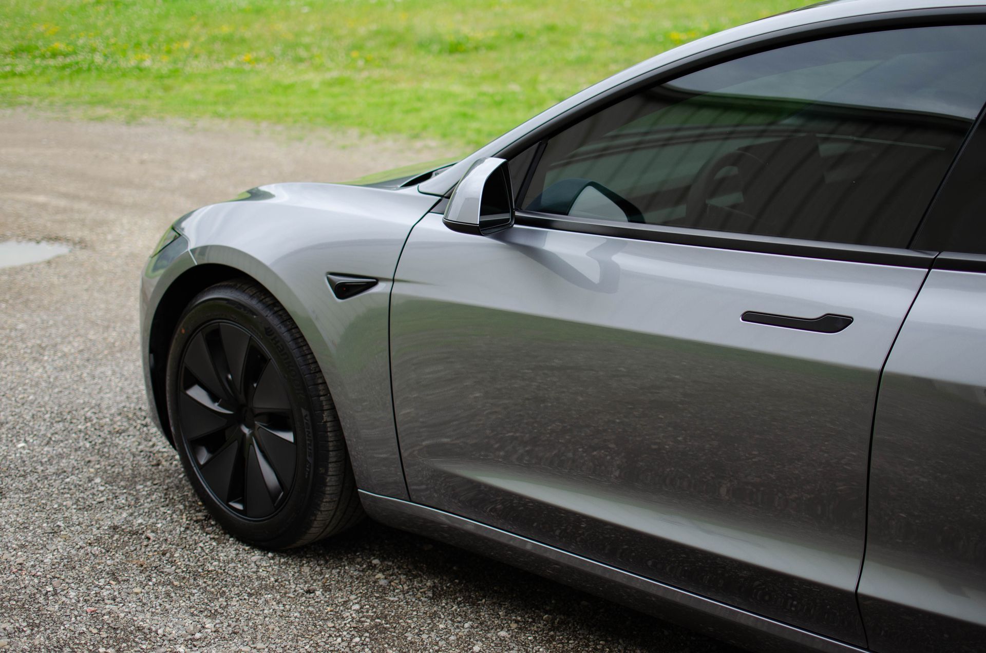 Silver Tesla sedan parked on a gravel driveway beside grass, front side view.