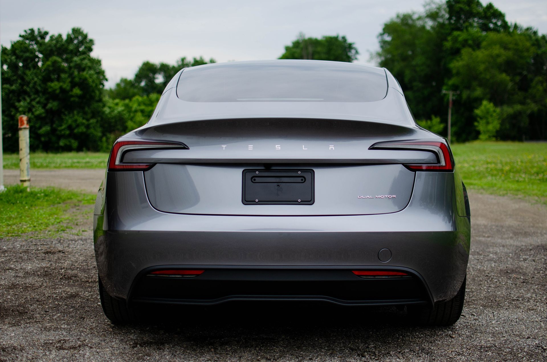 Rear view of a gray Tesla sedan parked on a gravel road with green trees and grass in the background