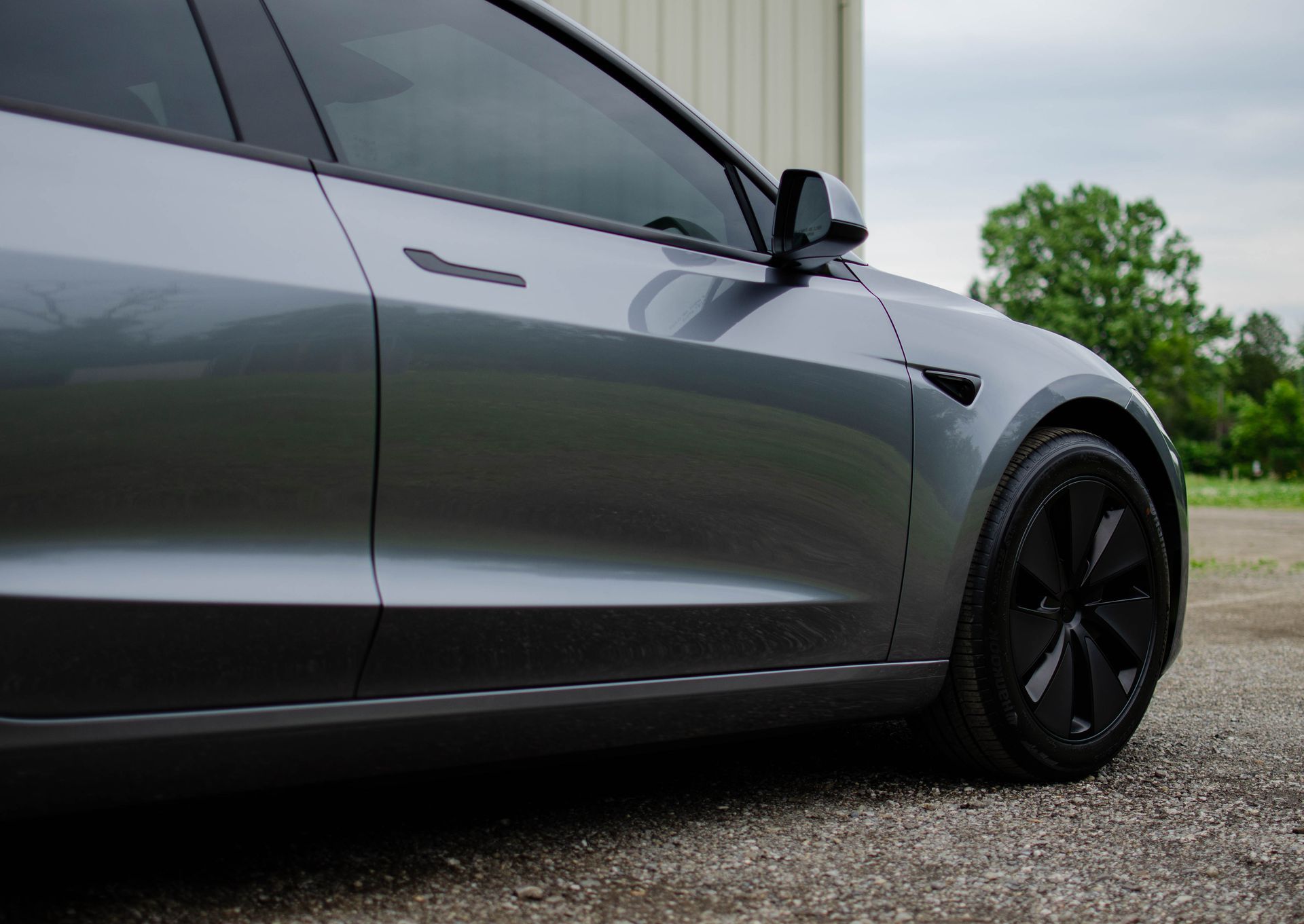 Gray sedan parked on gravel beside a building, showing the side and front wheel