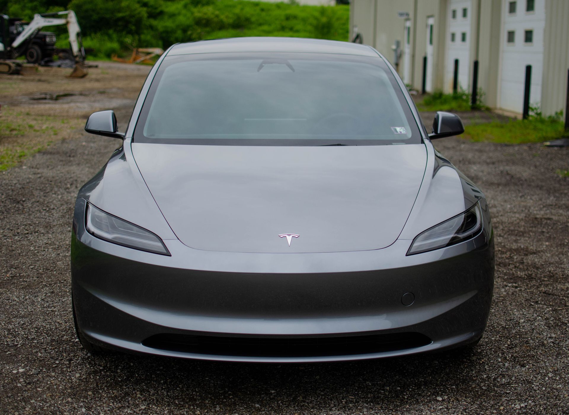 Front view of a gray Tesla parked on a gravel driveway beside a building.