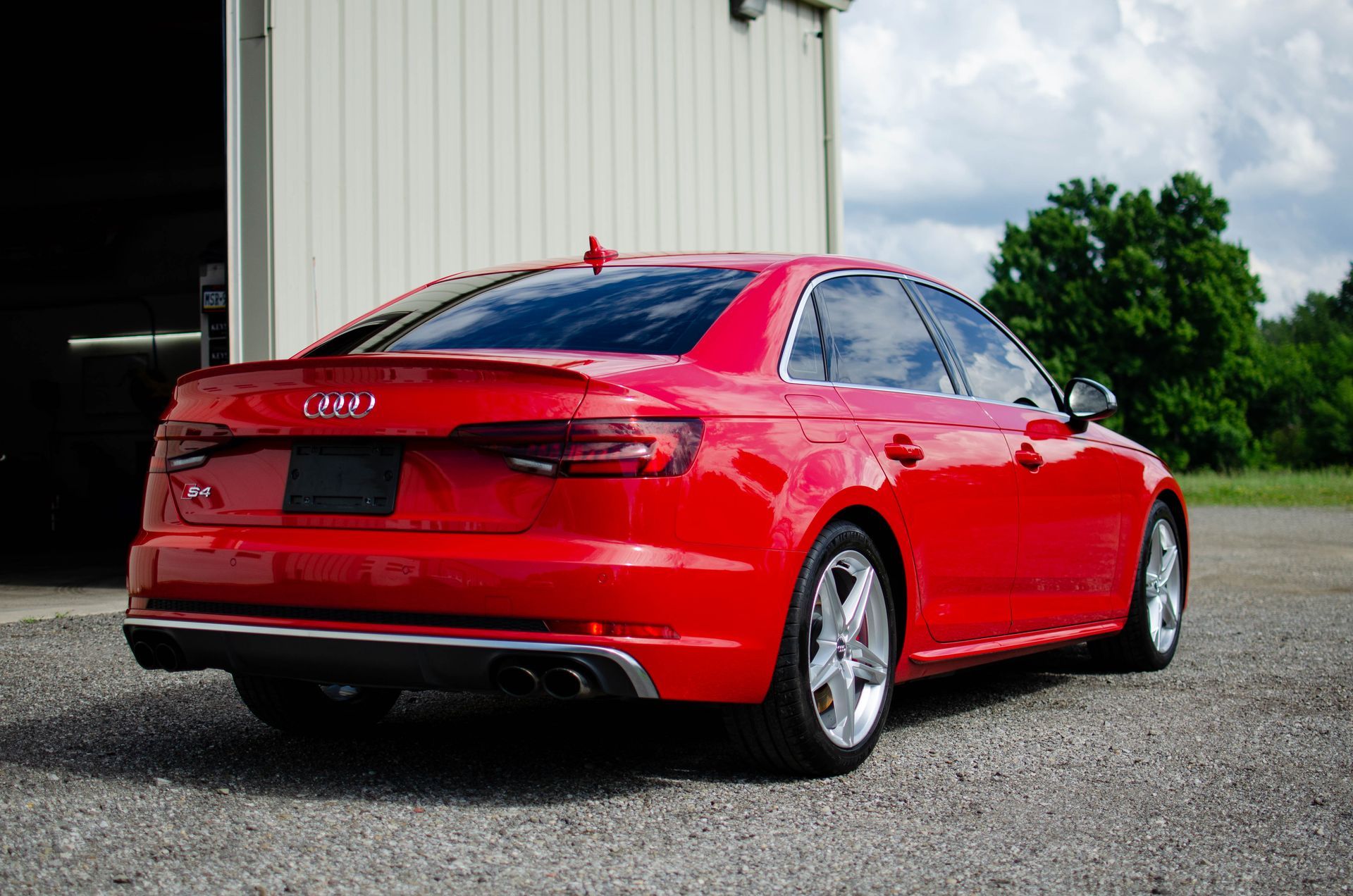 Red Audi sedan parked on gravel beside a white building, rear three-quarter view
