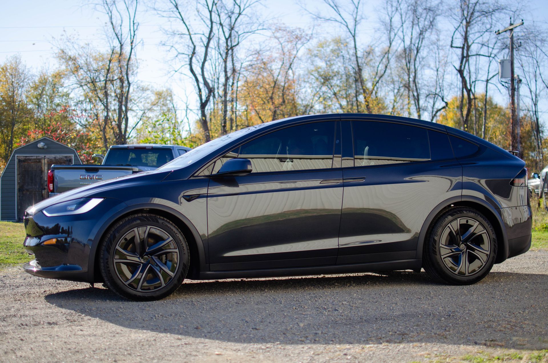 Dark blue Tesla Model Y parked on gravel in front of trees and a house at sunset