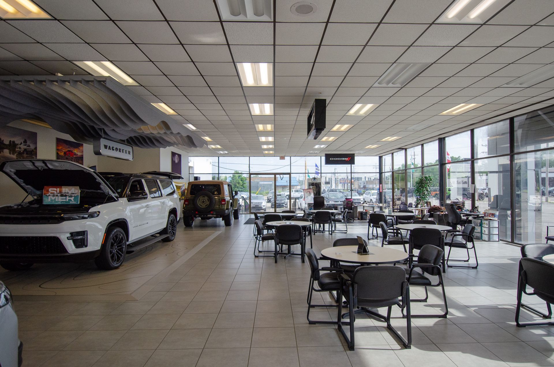 Car dealership showroom with white cars, tables, and large windows letting in daylight.