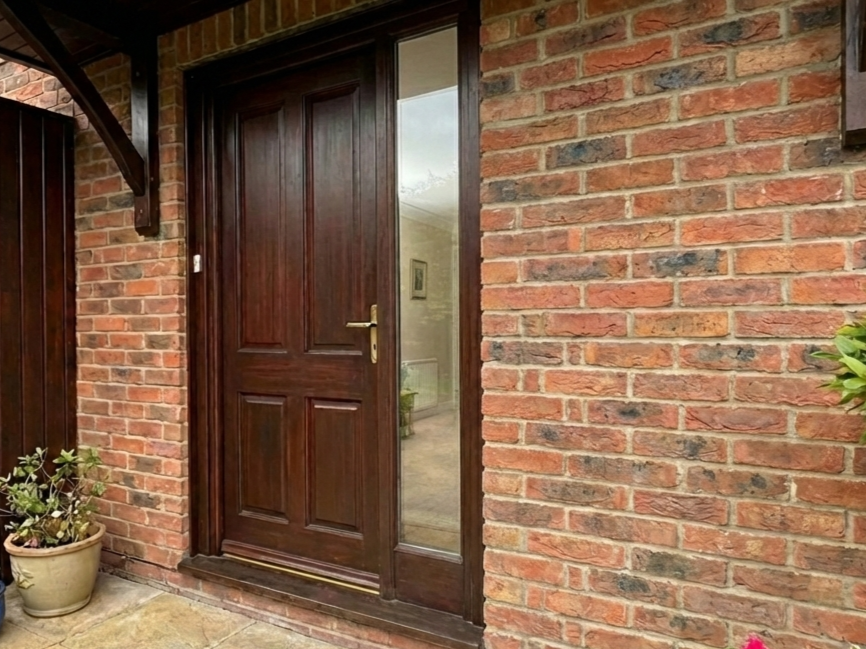 Brick house entrance with a dark wooden door, sidelight window, and potted plants on the left