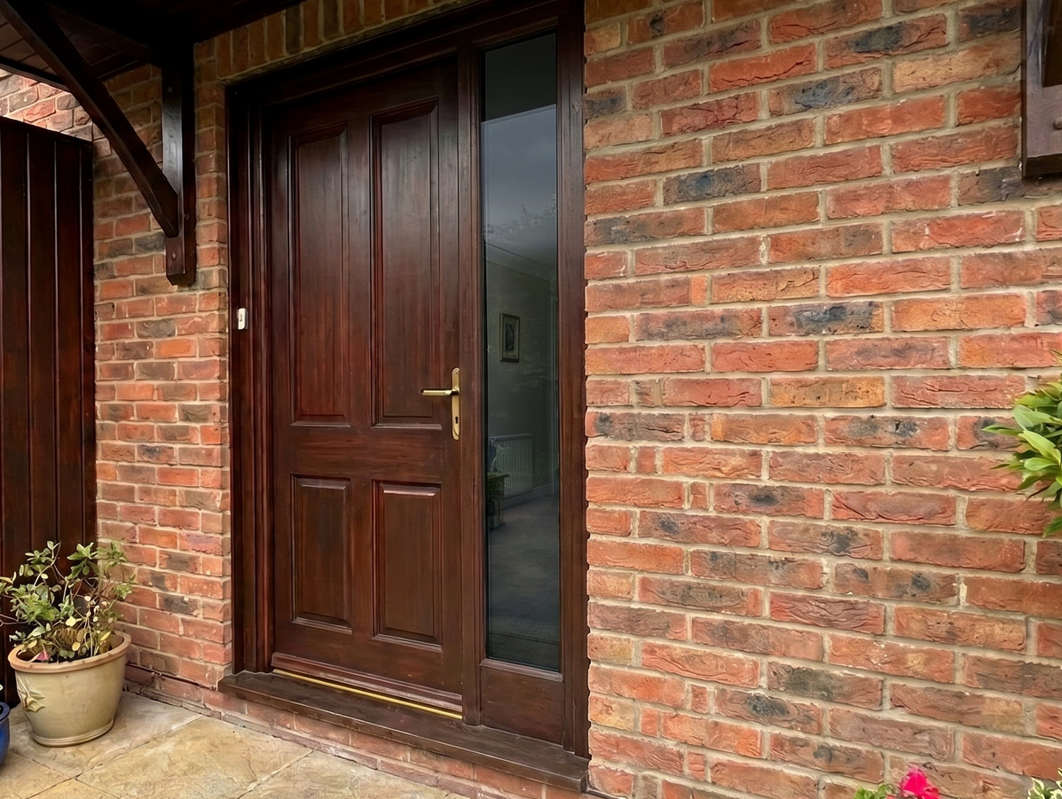 Brick house entry with a dark wooden front door and brick wall, flanked by a small potted plant.