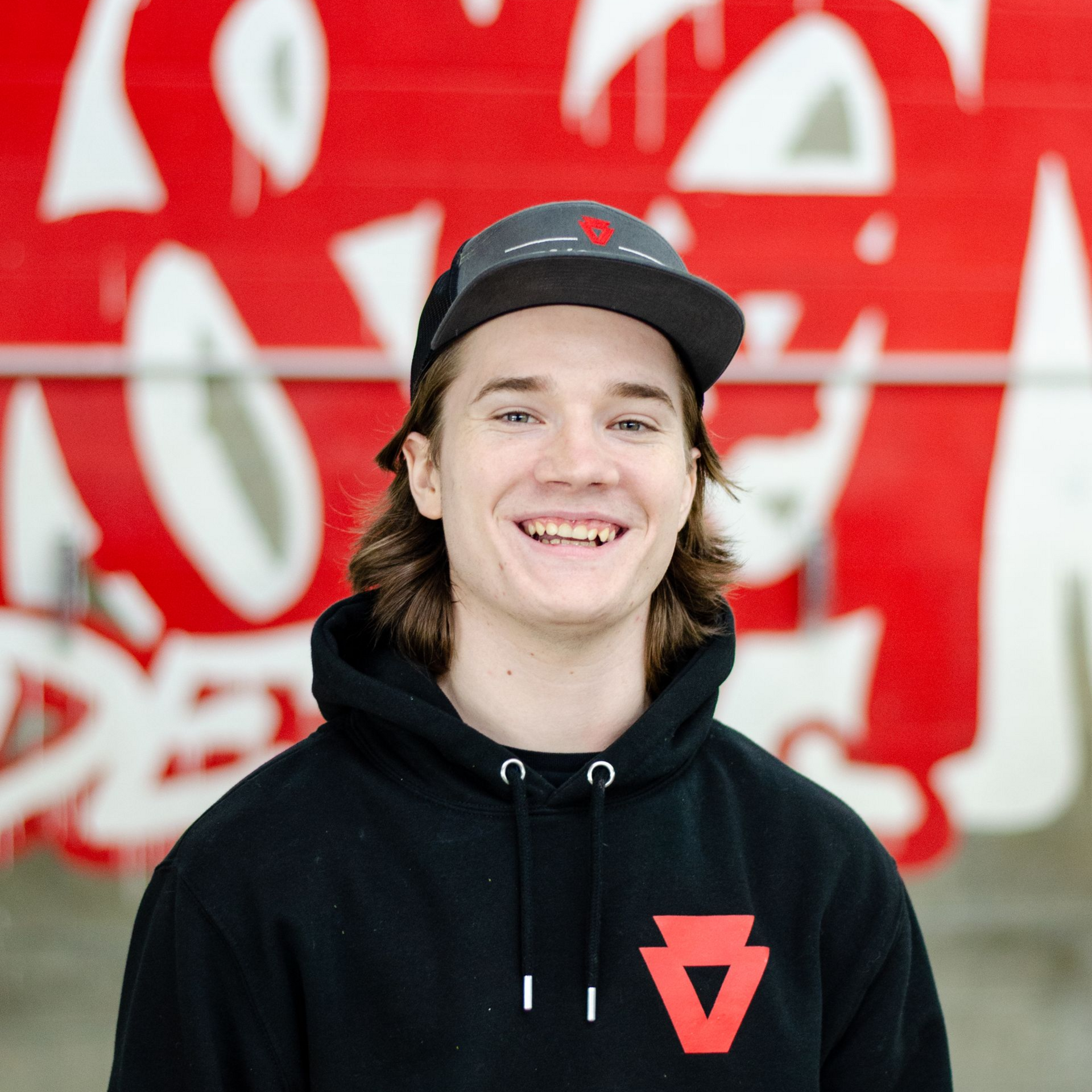 Smiling person in a black hoodie and cap in front of a red-and-white graffiti wall