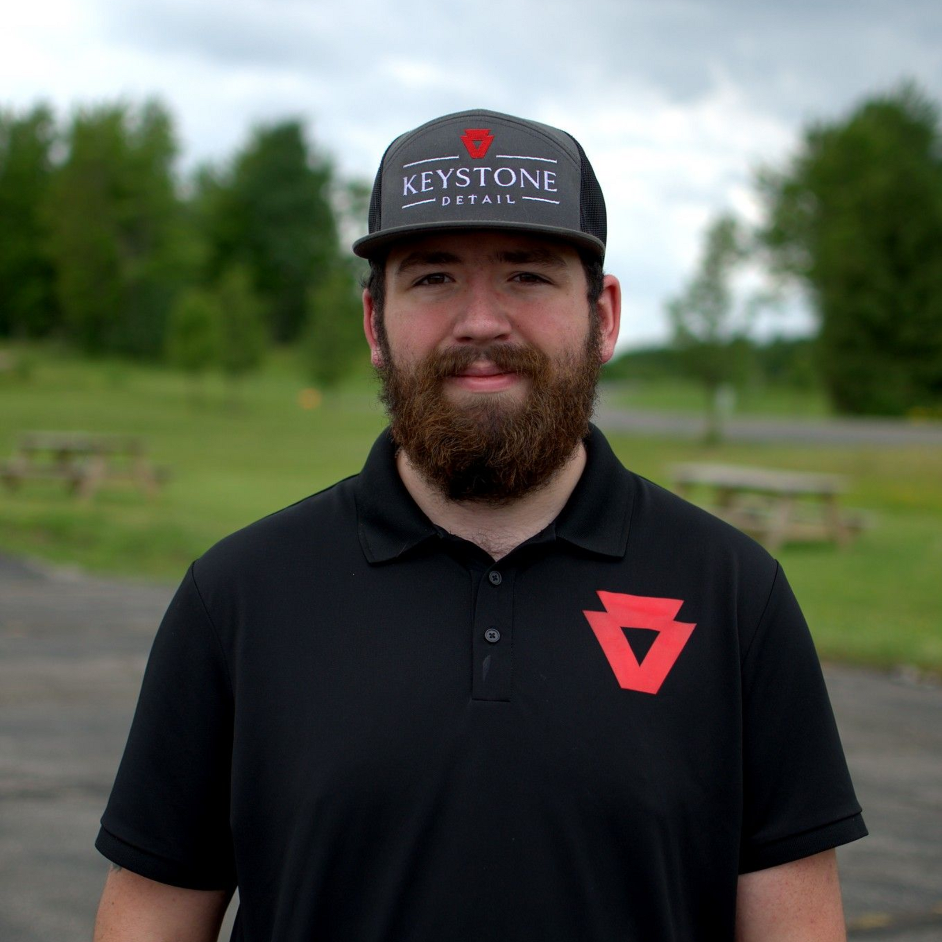 Bearded man in a black polo and Keystone cap standing outdoors on a grassy field.