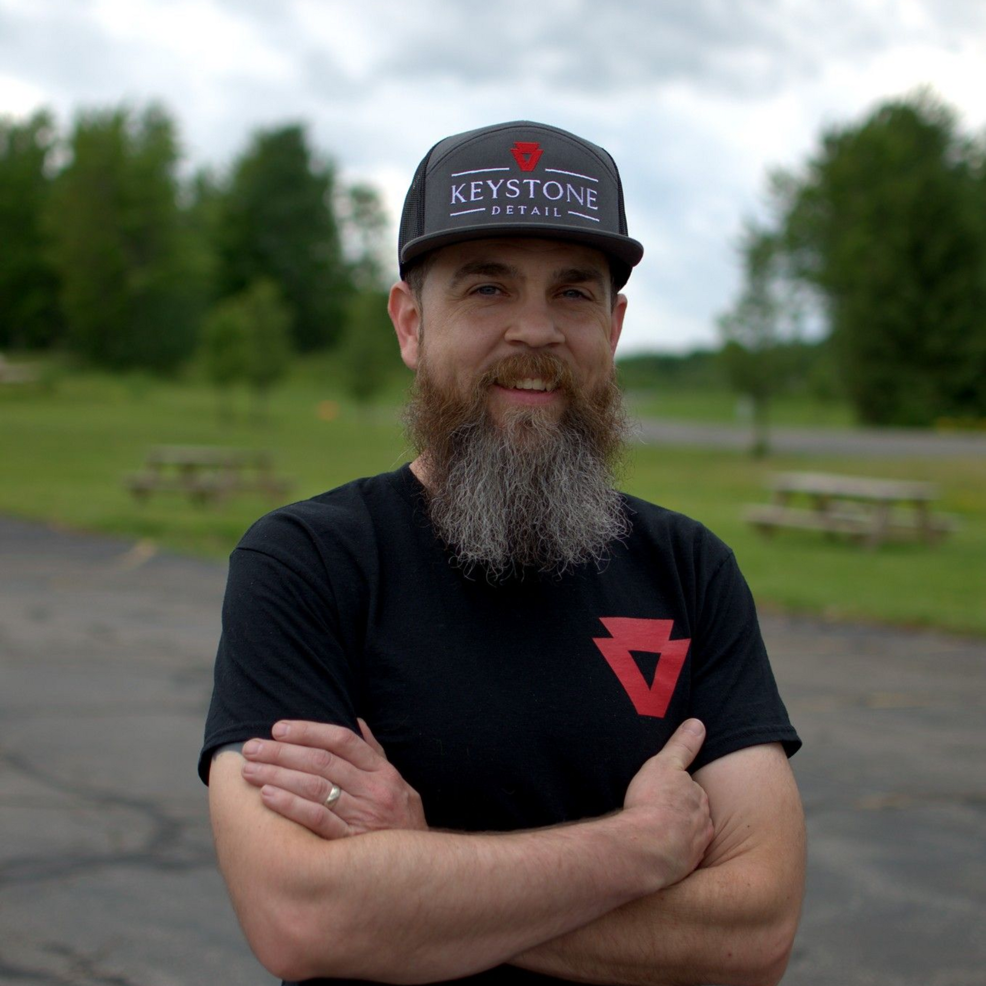 Bearded man in a black T-shirt and cap standing outdoors with arms crossed in a park.
