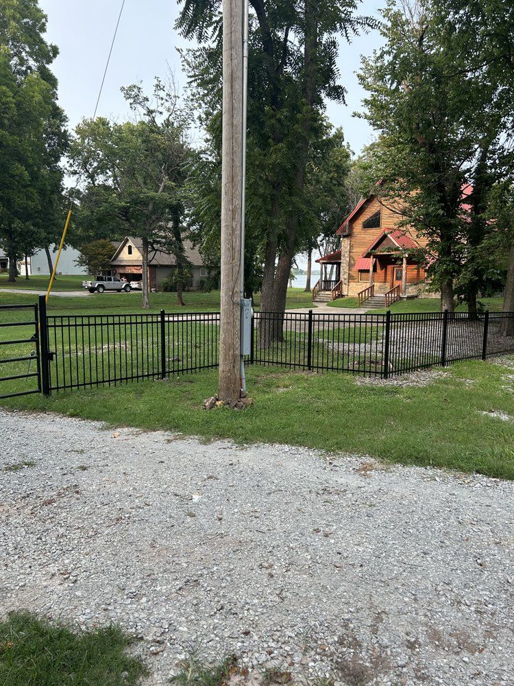 Gravel path and black fence in a wooded park, with two small wooden cabins in the background.