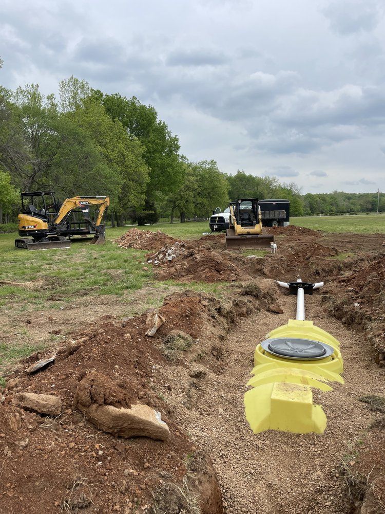 Trenching site with excavator, dump truck, and yellow utility fixture beside an open ditch under cloudy sky