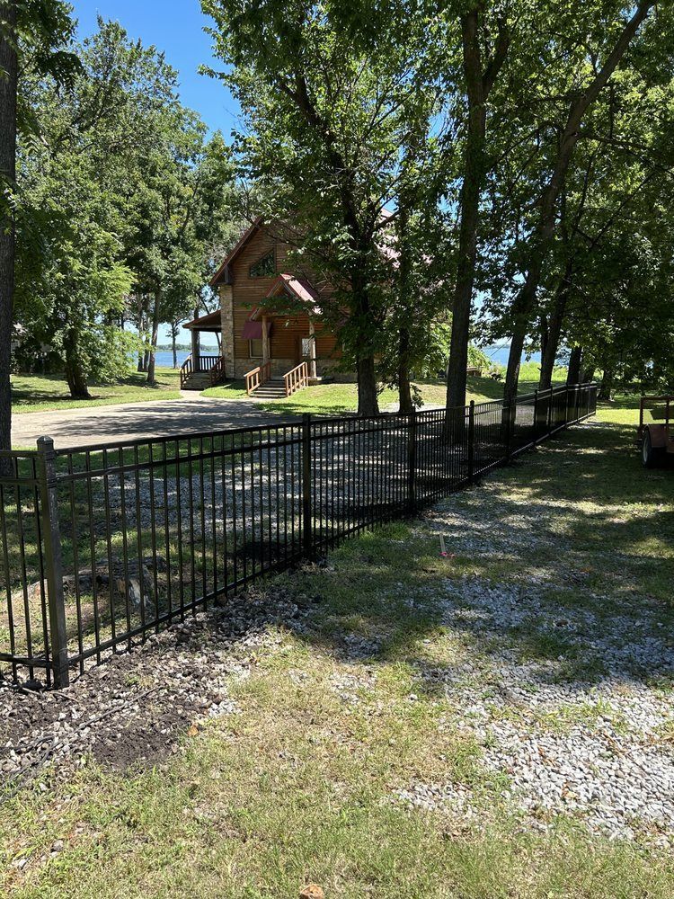 Black metal fence along a gravel path in a sunny wooded park with a small cabin in the background